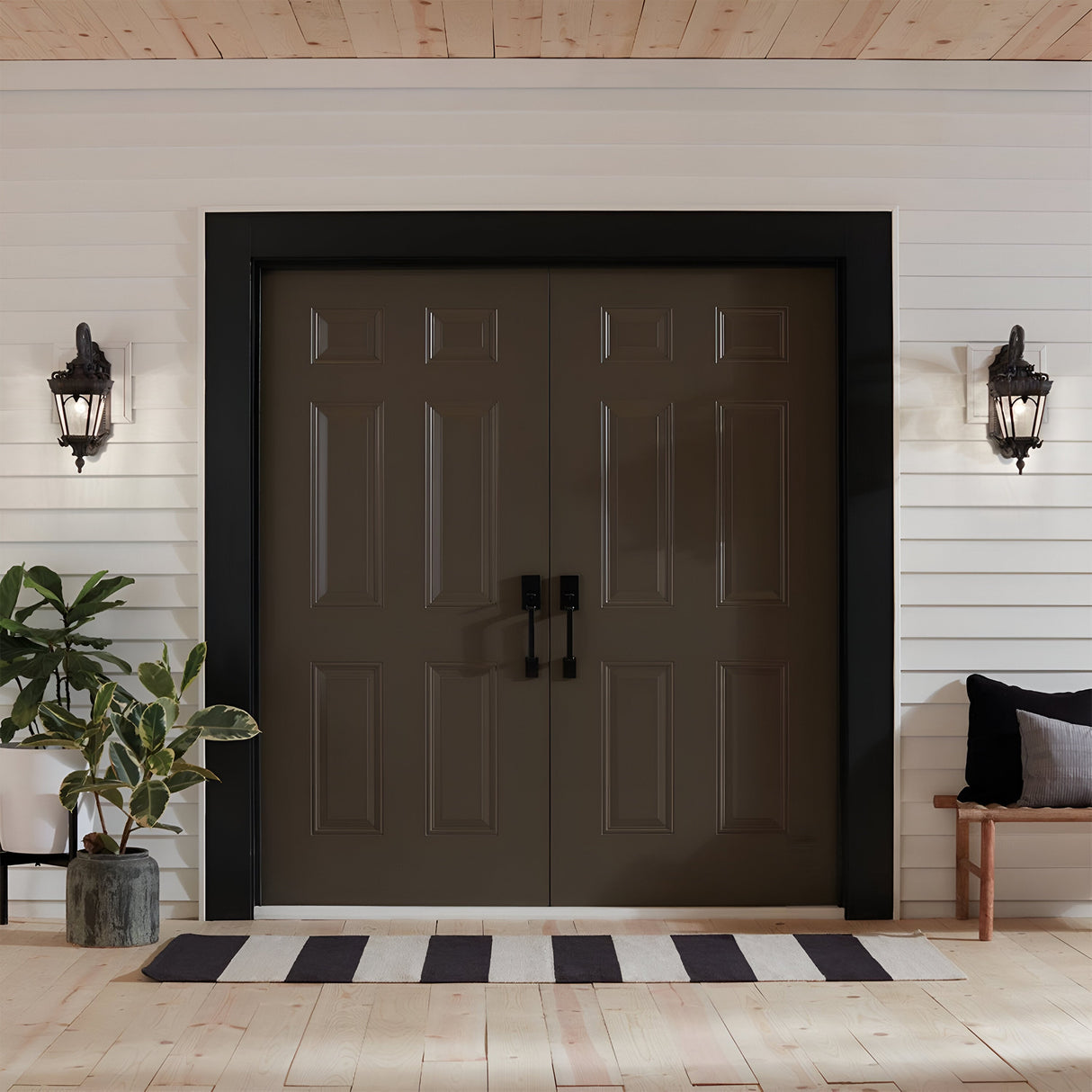 A pair of dark brown double doors with black handles is set in a white wall. Tournai Outdoor 2 Light Large Wall Lanterns in Bronze frame the entrance, showcasing a historic lantern design. To the right, a small bench topped with black and white pillows provides seating, while a potted plant adds greenery to the left, all beneath a wooden ceiling.