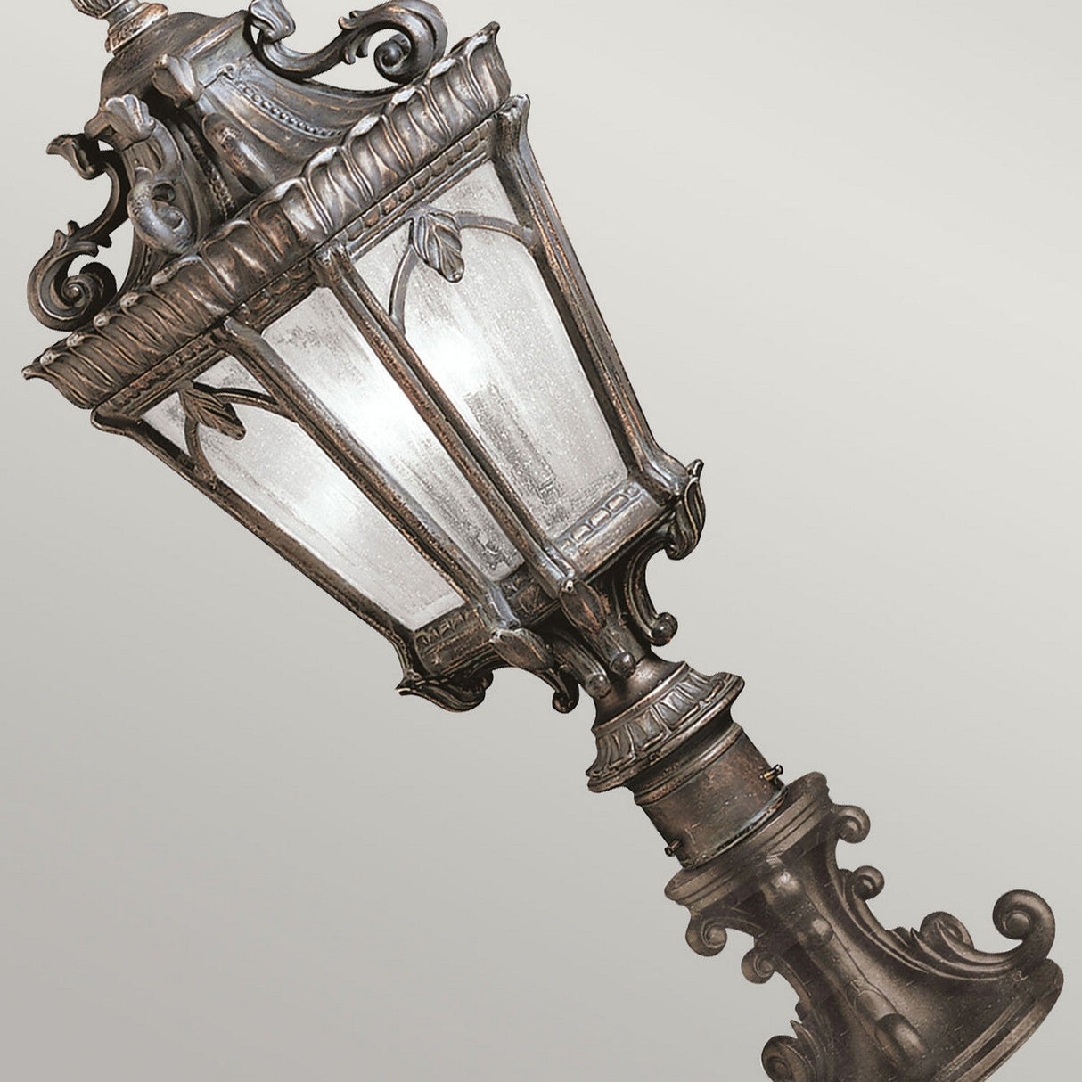 A Tournai 2 Light Large Pedestal in Bronze, featuring an ornate vintage-style street lamp design with a glass enclosure and decorative metalwork, complemented by clear seedy glass, shines against a light gray background.