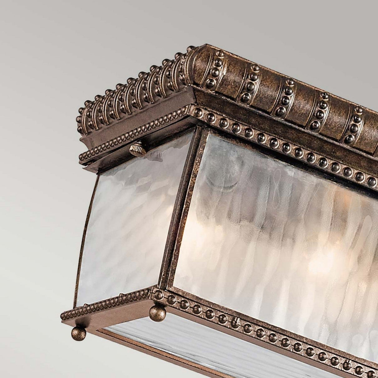 Close-up of an ornate, Renaissance design light fixture with a rectangular shape. The Venetian Rain Outdoor 2 Light Flush - Brushed Bronze features decorative brushed bronze metalwork and textured glass panels, emitting a soft glow while showcasing intricate beading and scrollwork details.