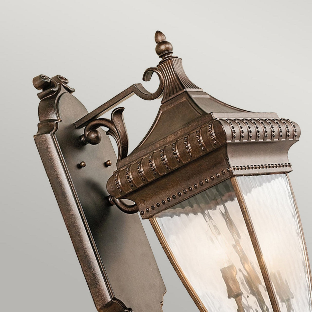 Close-up of the Venetian Rain Outdoor 3 Light Large Wall Lantern in brushed bronze, showcasing its decorative arm and glass panels mounted on a gray wall. The lantern's design highlights a textured surface with elegant detailing.