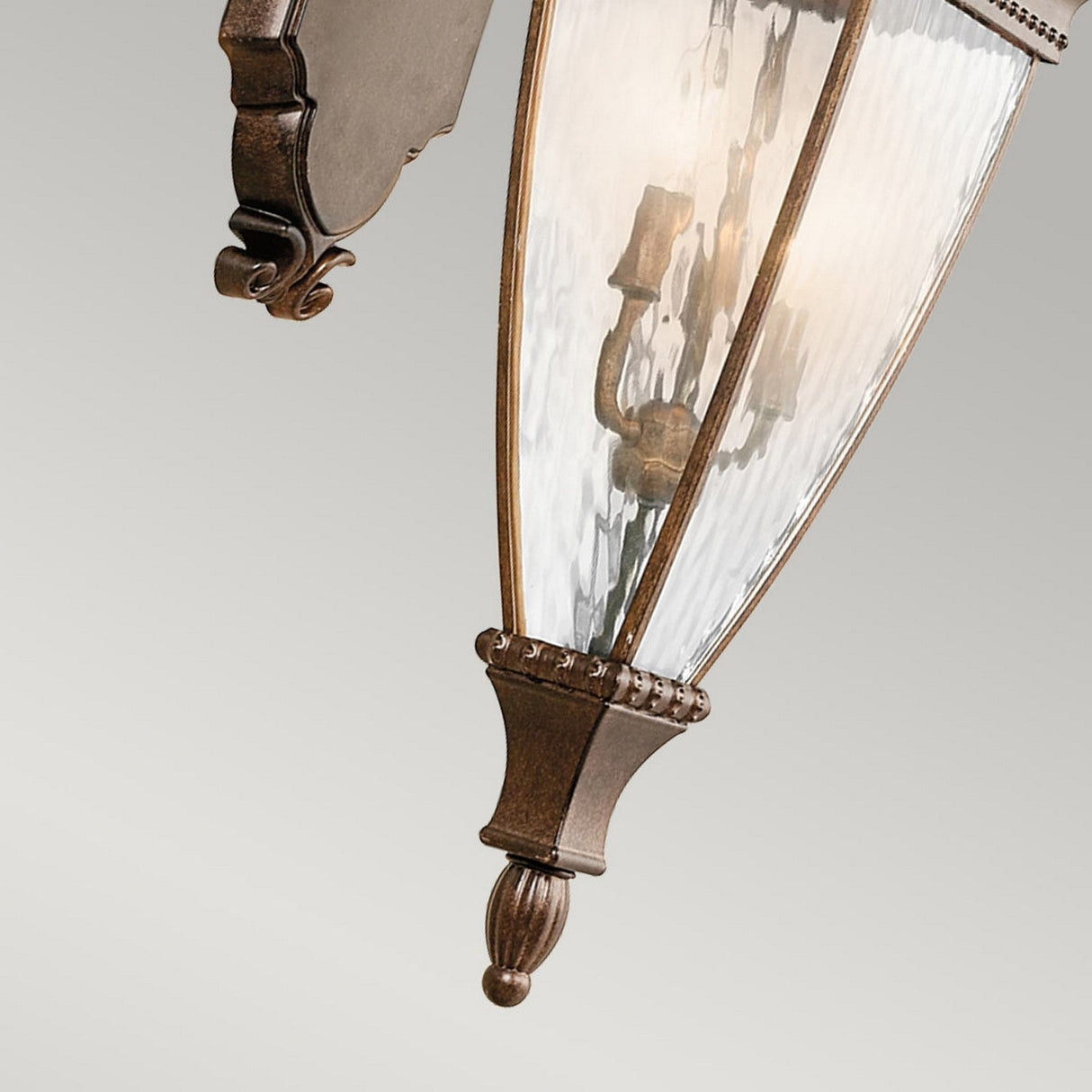 Close-up of the elegant Venetian Rain Outdoor 3 Light Large Wall Lantern, showcasing a glass enclosure with a brushed bronze finish and intricate decorative detailing. It houses three candle-style bulbs inside, casting a warm glow with its vintage and ornate design.