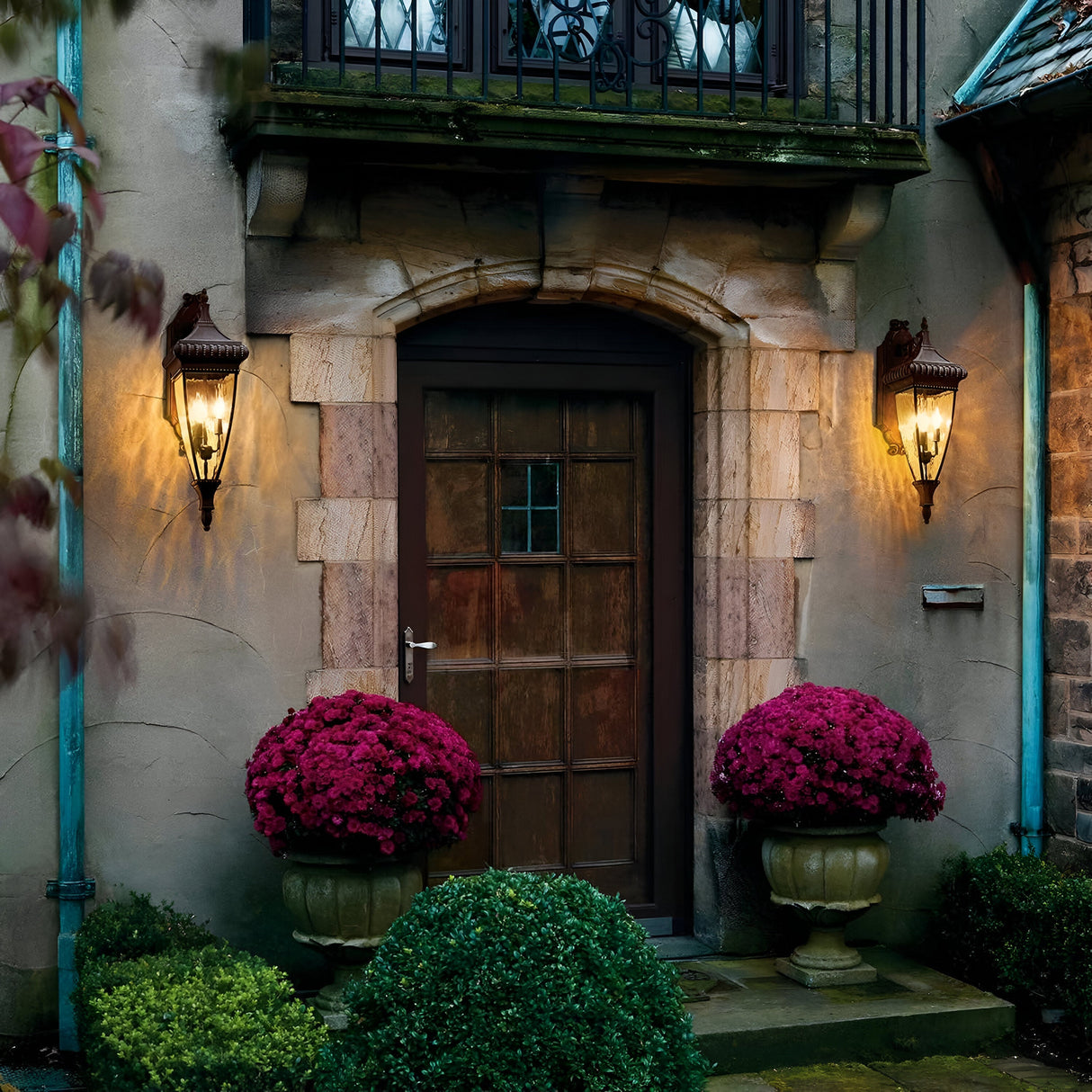 A rustic stone building entrance with a wooden door is flanked by the glowing Venetian Rain Outdoor 3 Light Large Wall Lanterns in brushed bronze. Large pots overflowing with vibrant purple flowers sit amidst green shrubs. Above, a small balcony features an ornate railing that complements the brushed bronze accents of the scene.