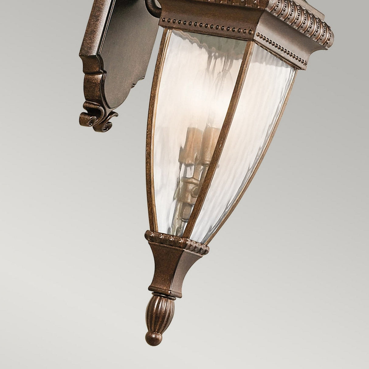 A close-up of the Venetian Rain Outdoor 2 Light Medium Wall Lantern - Brushed Bronze highlights its Italian design with a brushed bronze metal frame and clear, curved glass panels. This vintage-style lantern, designed for wall mounting, features two candle-like bulbs against a plain neutral backdrop.
