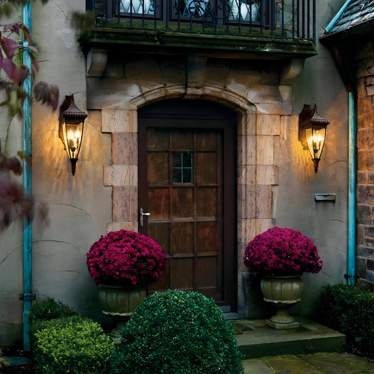 A rustic wooden door set in a stone entryway is flanked by two Venetian Rain Outdoor 2 Light Medium Wall Lanterns in brushed bronze and symmetrical planters filled with lush pink flowers. Trimmed green bushes line the pathway, enhancing the welcoming ambiance of the Italian design.
