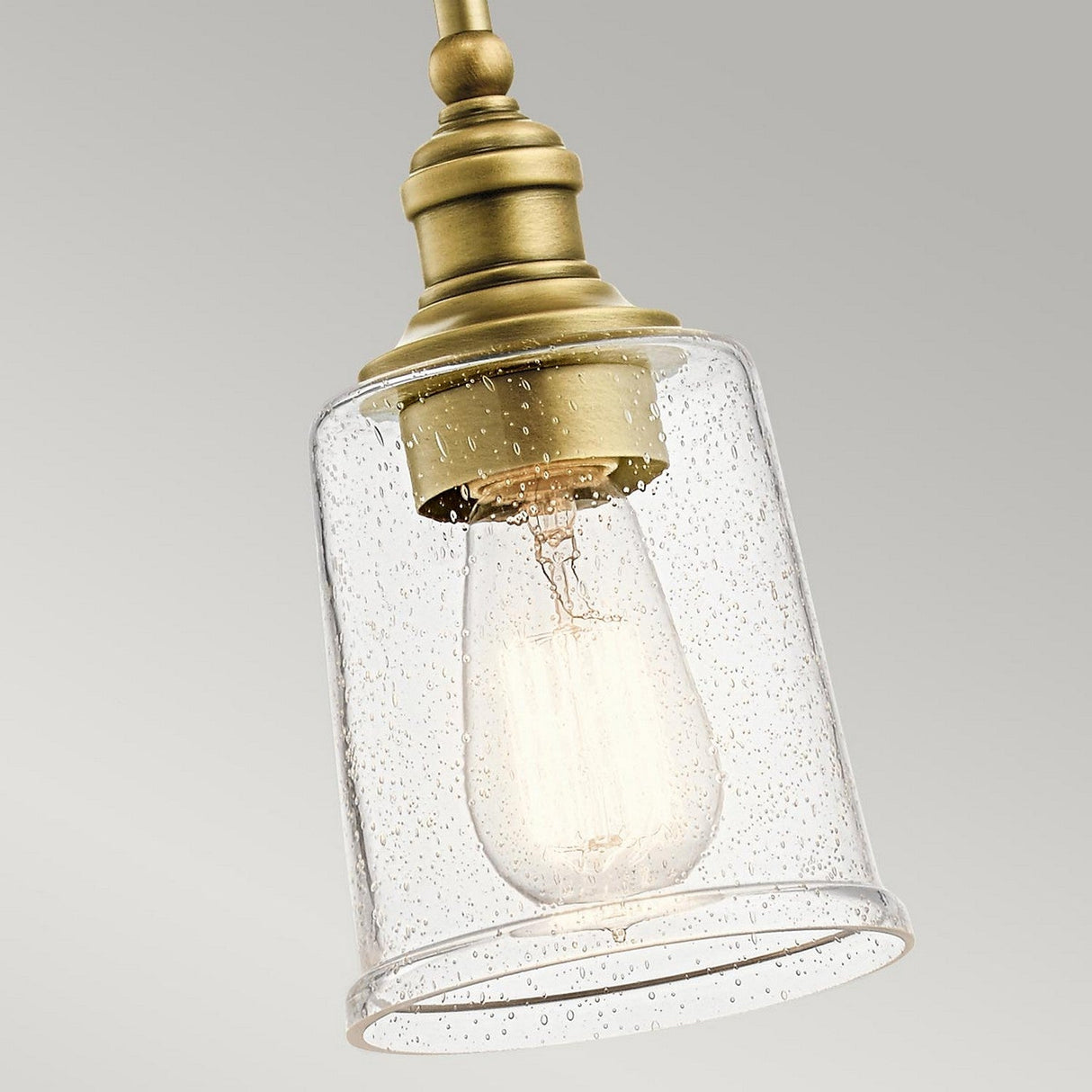 Introducing the Waverly 1 Light Mini Pendant in Natural Brass: This vintage-inspired fixture boasts a clear glass shade adorned with delicate bubbles. The Edison-style bulb inside emits a warm glow, contrasting beautifully with a neutral gray backdrop.