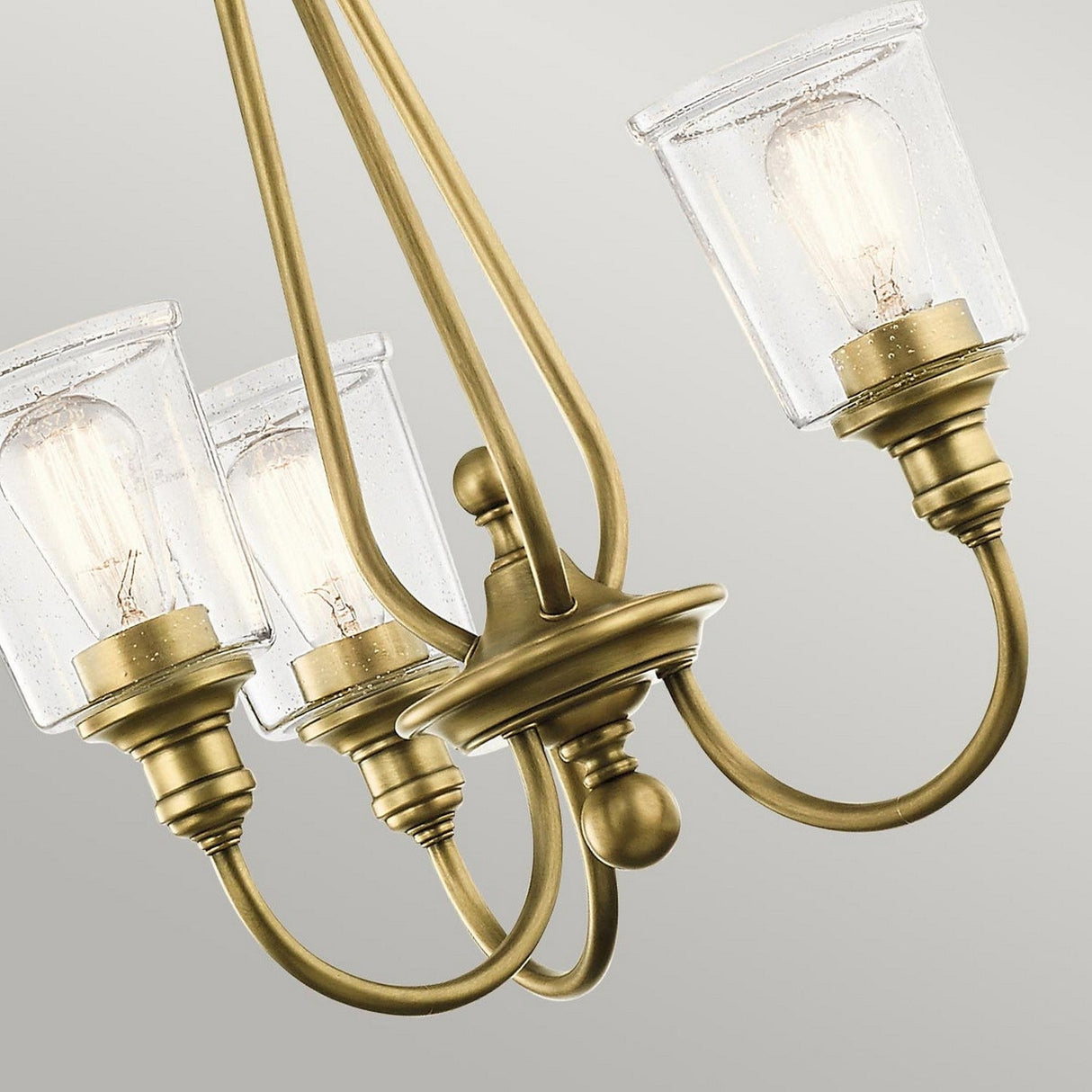 A close-up of the Waverly 3 Light Chandelier in natural brass showcases its three elegantly curved arms, each featuring a seeded glass shade and a vintage-style light bulb. The chandelier radiates a sense of vintage elegance against a plain gray background.