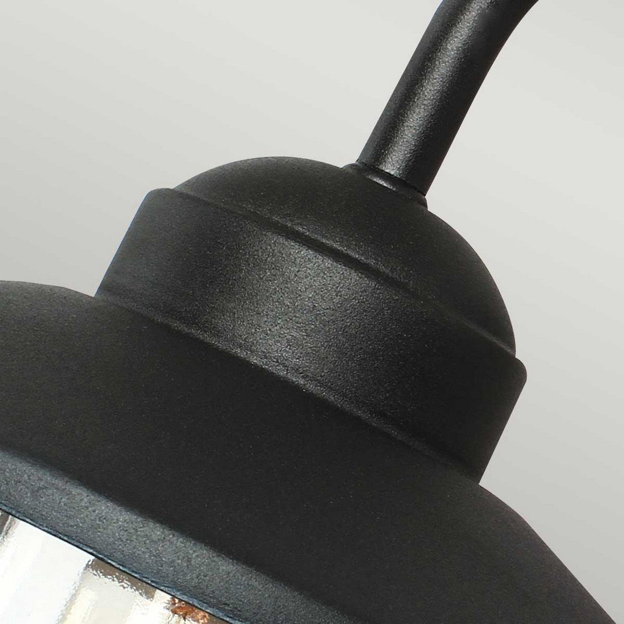 Close-up of the Klampenborg 1 Light Outdoor Wall Light in black, showcasing an industrial-style design with a curved shade that embraces Scandinavian aesthetics. The matte, textured surface highlights a visible cylindrical stem at the top, partially enveloping the glass around the bottom edge.