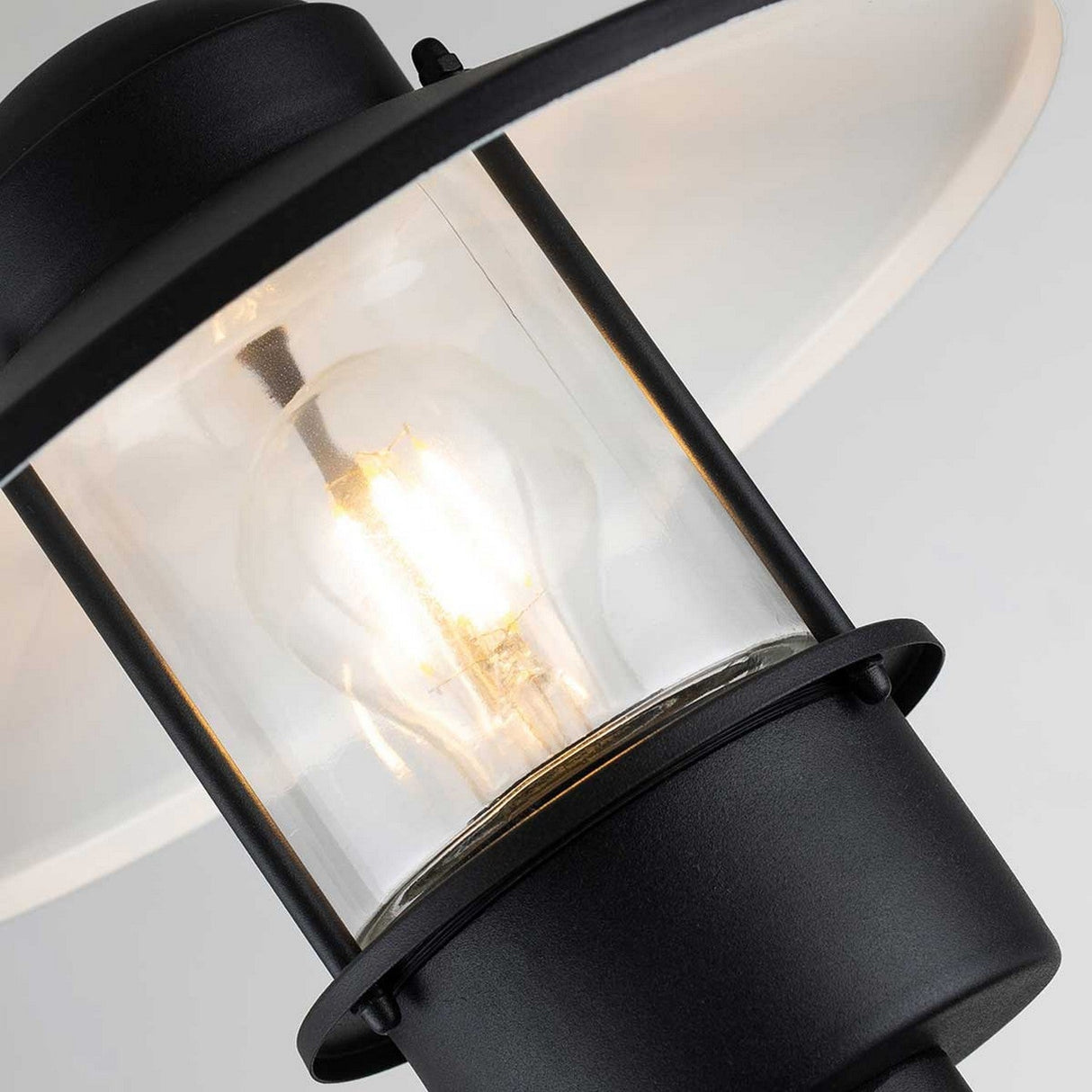 Close-up of a black outdoor lantern with a clear glass enclosure, revealing a glowing light bulb within. The lamp is showcased against a neutral background, highlighting its simple yet elegant design, ideal for enhancing any outdoor lighting arrangement alongside the Klampenborg 1 Light Bollard - Black.