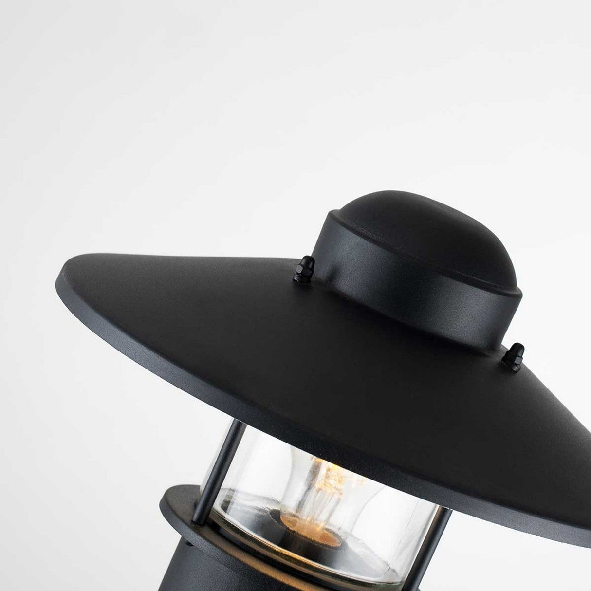 Close-up of the Klampenborg 1 Light Bollard in black, featuring a modern design with a wide, flat top and a clear glass enclosure that showcases a visible light bulb. The plain white wall background highlights its contemporary elegance.