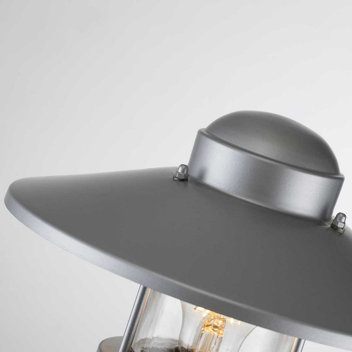 A detailed view of the Klampenborg 1 Light Bollard in silver showcases its gray metal structure with a rounded cap and transparent glass covering, partially exposing the bulb inside. This corrosion-resistant fixture features a modern, minimalist Scandinavian design set against a neutral background.
