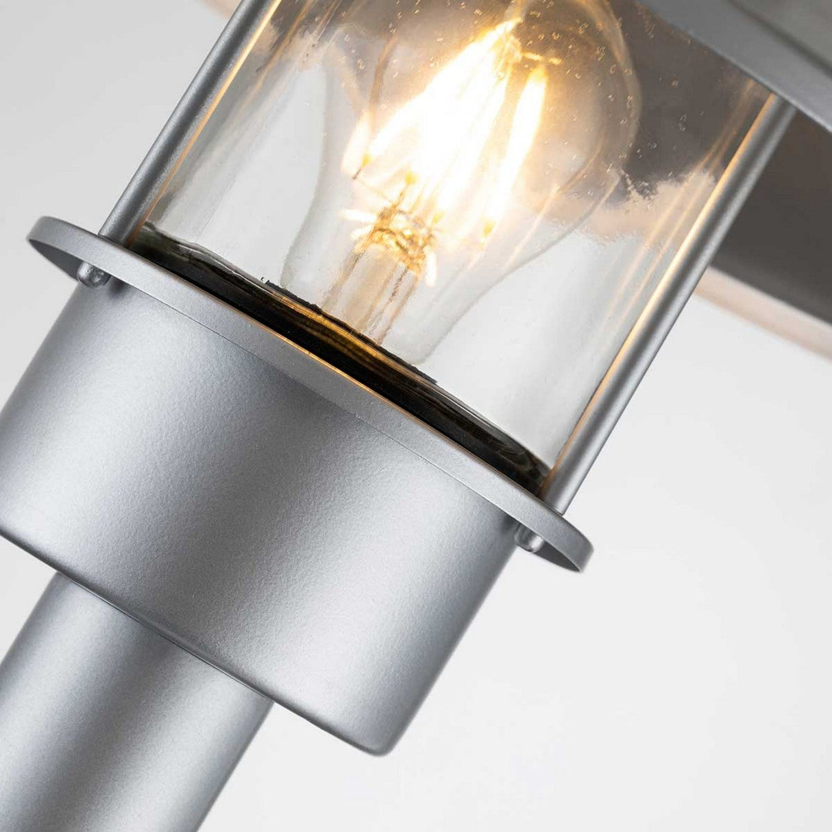 A close-up view of the Klampenborg 1 Light Bollard in silver reveals its modern, corrosion-resistant metal construction with a clear glass cover. The illuminated bulb inside emits a warm glow, accentuating the sleek Scandinavian design of the fixture.