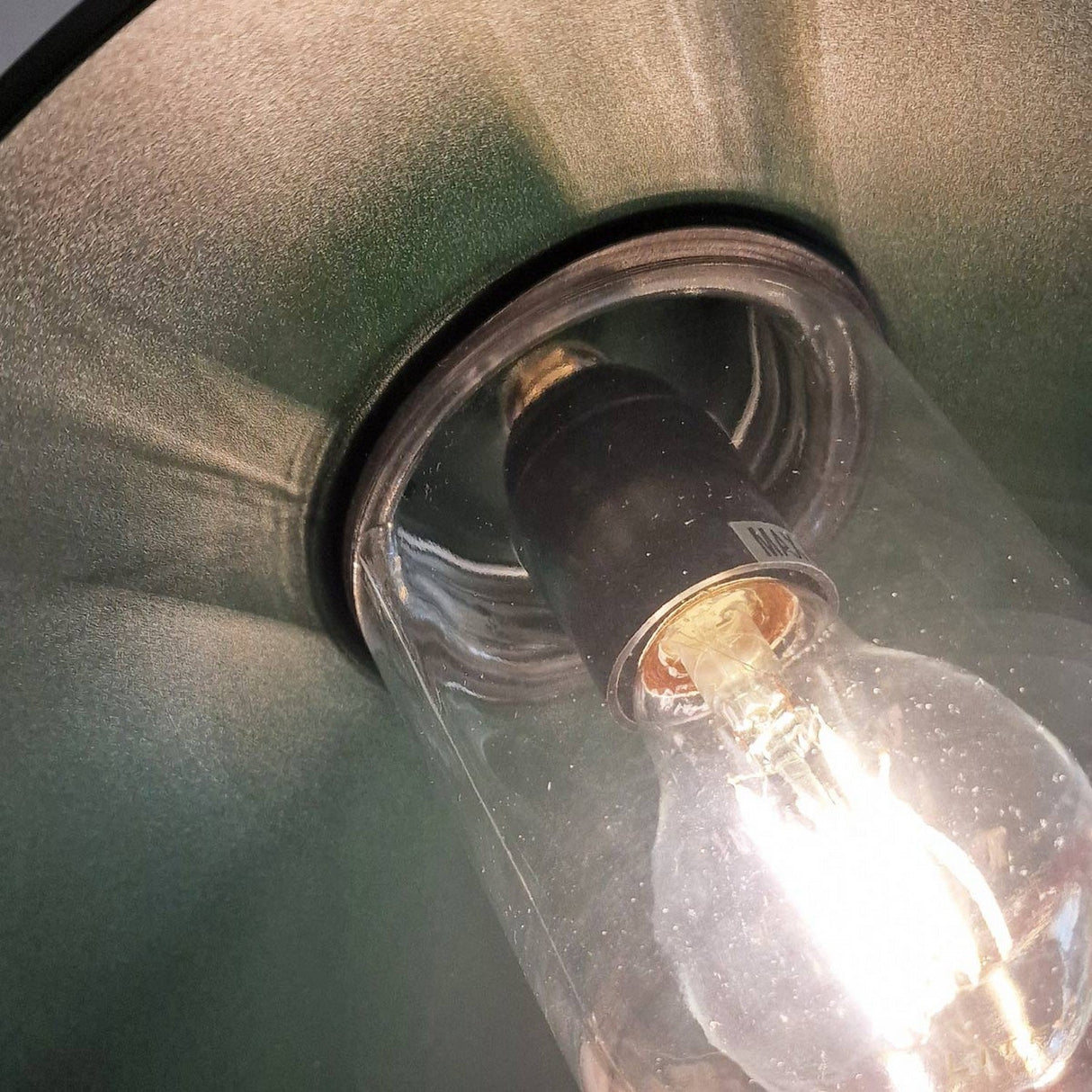 A close-up of the lit Klampenborg 1 Light Outdoor Wall Light in green showcases an incandescent bulb emitting a warm glow, casting intricate patterns of light and shadow on the inner rim. The Scandinavian design features a protective glass casing and a metal base that are also visible.