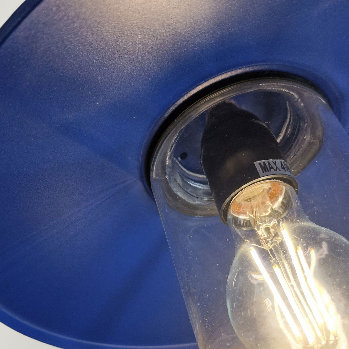 A close-up of the Klampenborg 1 Light Outdoor Wall Light in navy blue highlights its Scandinavian-inspired design. The visible filaments in the bulb emit a warm glow, and the fixture's label indicates a maximum wattage of 40W. The lampshade boasts a shiny, metallic finish that enhances its modern aesthetic.