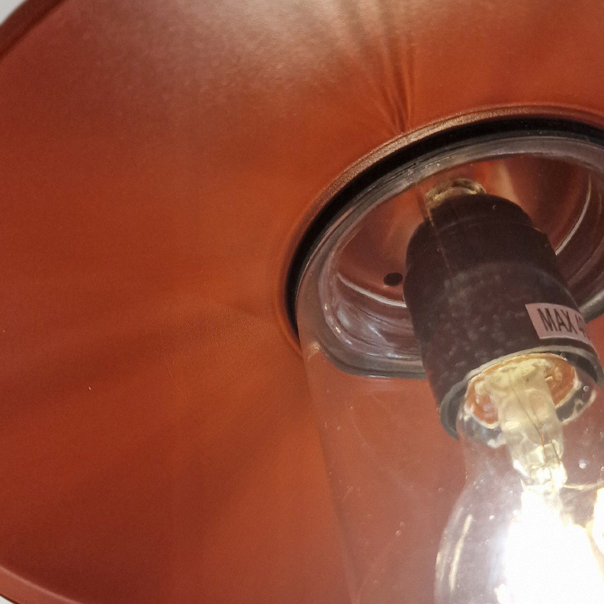 A close-up view of the Klampenborg 1 Light Outdoor Wall Light in oxide red shows an illuminated light bulb with a visible filament inside its Scandinavian-style lamp design. It's surrounded by an oxide red lampshade, and a small label is attached, while the light casts a warm glow.