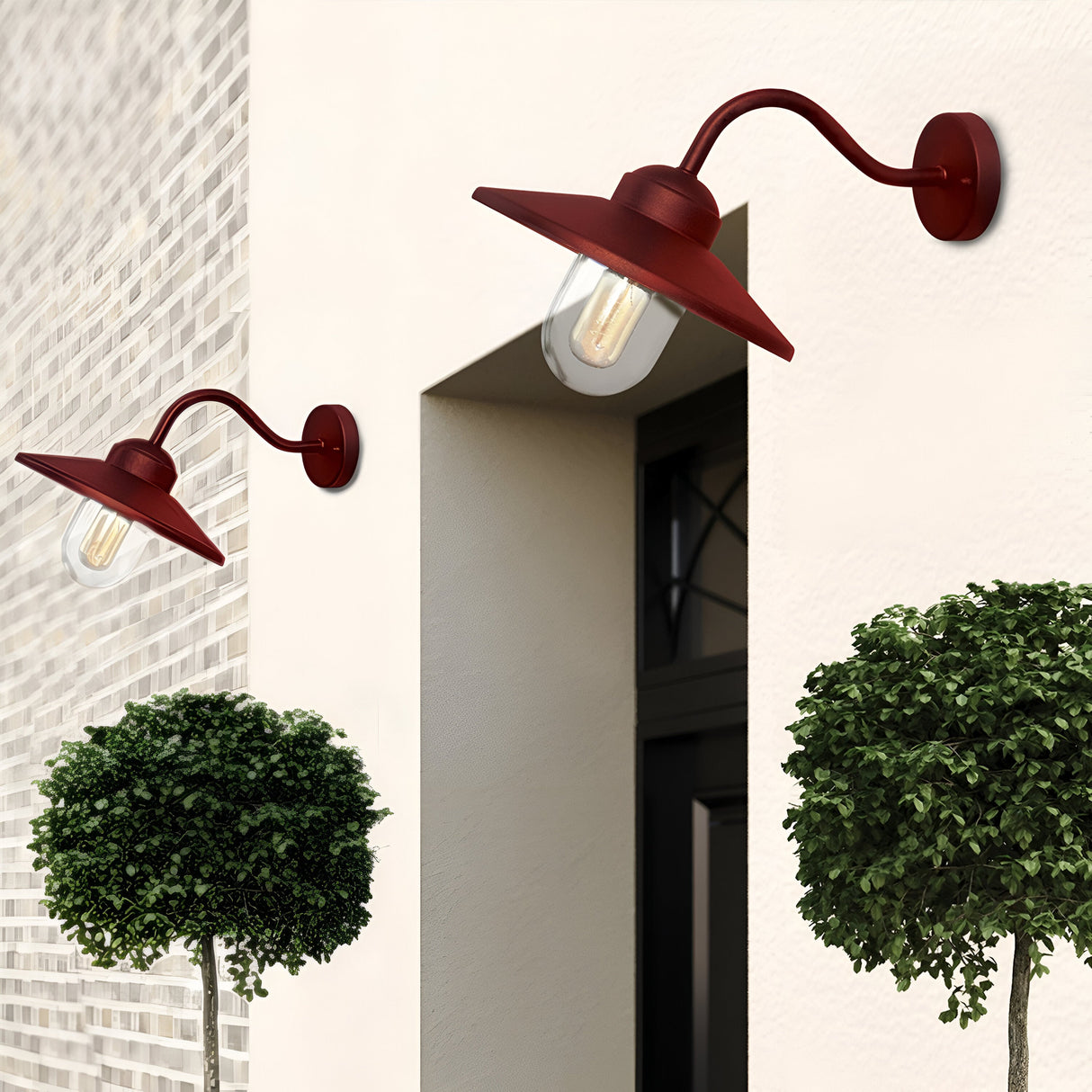 A pair of Klampenborg 1 Light Outdoor Wall Lights in an Oxide Red finish with clear bulbs adorn a beige wall. They elegantly frame a modern doorway and are accompanied by neatly pruned round bushes, adding a touch of Scandinavian style charm to the entrance.