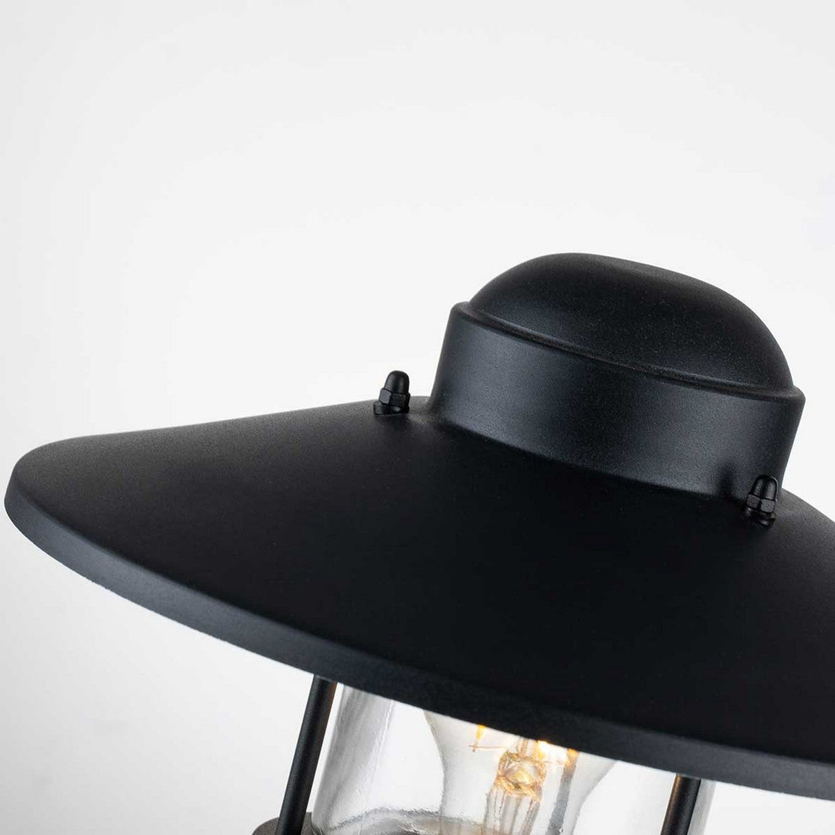 Close-up of the Klampenborg 1 Light Pedestal - Black, showcasing its Scandinavian design with a round top and a wide, slightly sloped shade. The lantern is illuminated, casting a warm glow through the clear glass encasement, set against a plain white background.