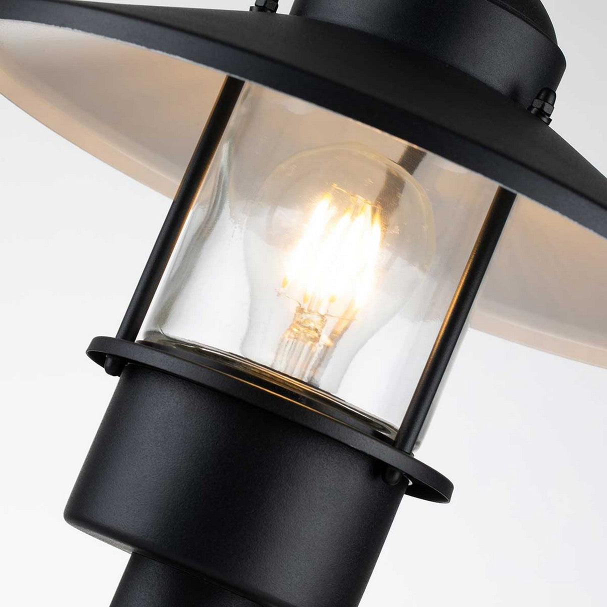 A close-up of the Klampenborg 1 Light Pedestal - Black, a Scandinavian design black metal lantern with clear glass, displays a glowing bulb inside. It features a wide-brimmed top and is set against a plain white background, making it perfect for enhancing your outdoor lighting ambiance.