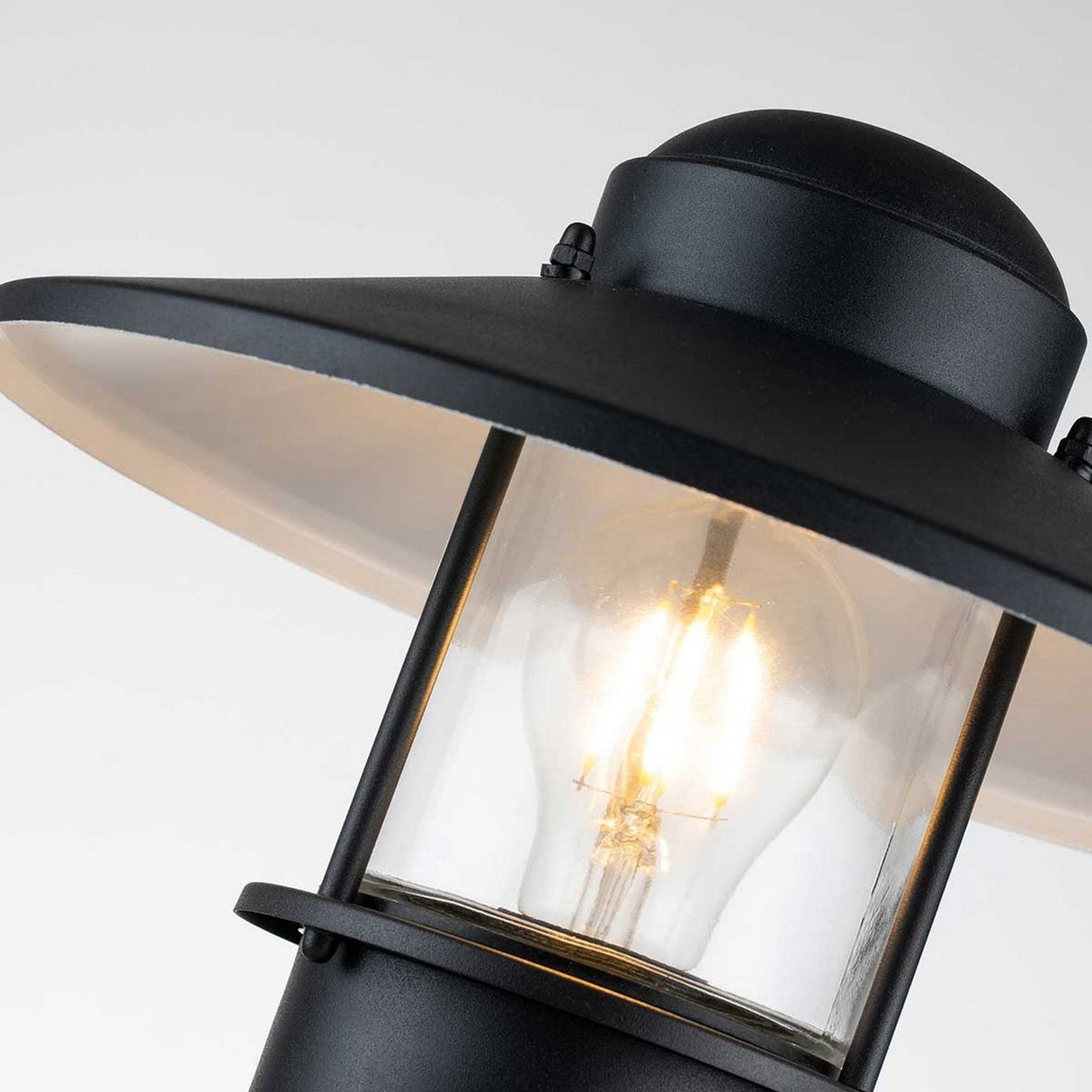 Close-up of the Klampenborg 1 Light Pedestal with PIR in black, an outdoor security lantern showcasing a glowing bulb inside. It features a wide circular shade and clear glass panels, set against a soft gray background.