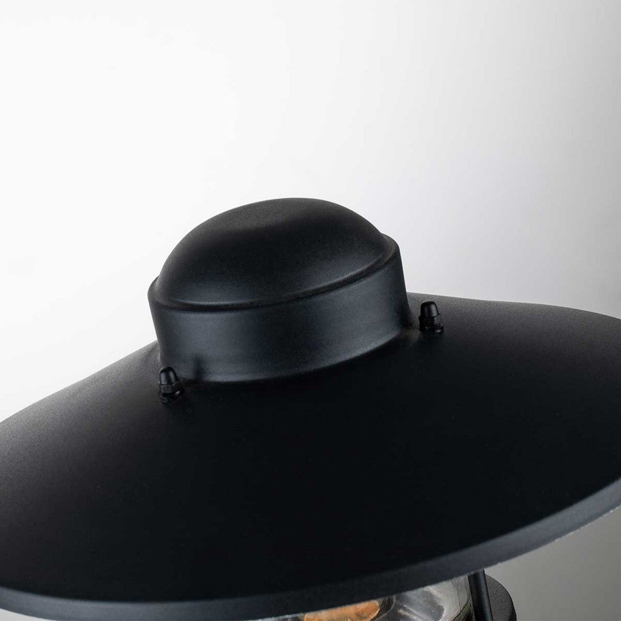 The Klampenborg 1 Light Pedestal with PIR - Black showcases a black metal outdoor light fixture featuring a dome-shaped top and a wide, flat shade, enhanced by an integrated PIR sensor for additional security against a plain light grey setting.