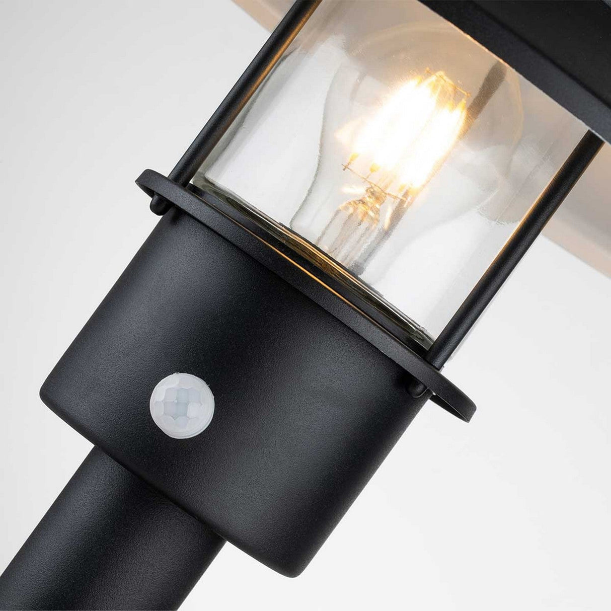 A close-up view of the Klampenborg 1 Light Pedestal with PIR - Black shows a glowing bulb inside its glass casing. This black outdoor security lantern includes a PIR sensor at its base for enhanced safety, set against a light gray background.