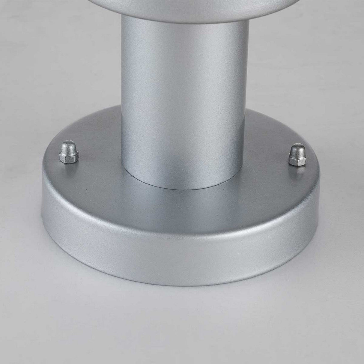 The Klampenborg 1 Light Pedestal with PIR - Silver features a sleek metallic cylinder design with a round base, secured by four bolts. Its smooth, silver finish gleams in the light, evoking the elegance of Scandinavian design. Perfect for outdoor lighting solutions, its neutral gray background complements its modern aesthetic effortlessly.
