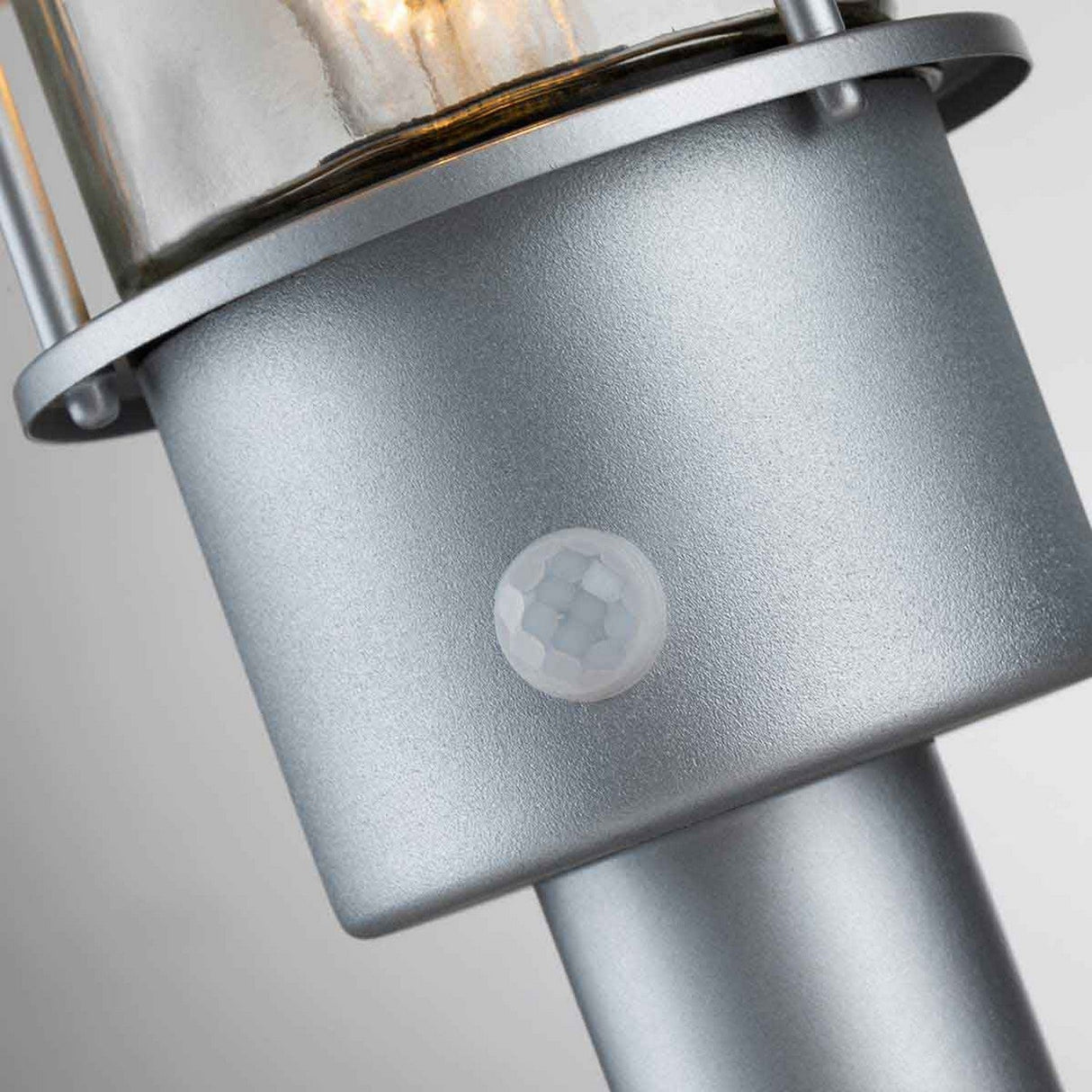 A detailed view of the Klampenborg 1 Light Pedestal with PIR - Silver highlights its Scandinavian design, showcasing a visible bulb and round PIR sensor at the base against a neutral backdrop. The pedestal features a cylindrical shape with a smooth finish, offering sleek outdoor lighting aesthetics.