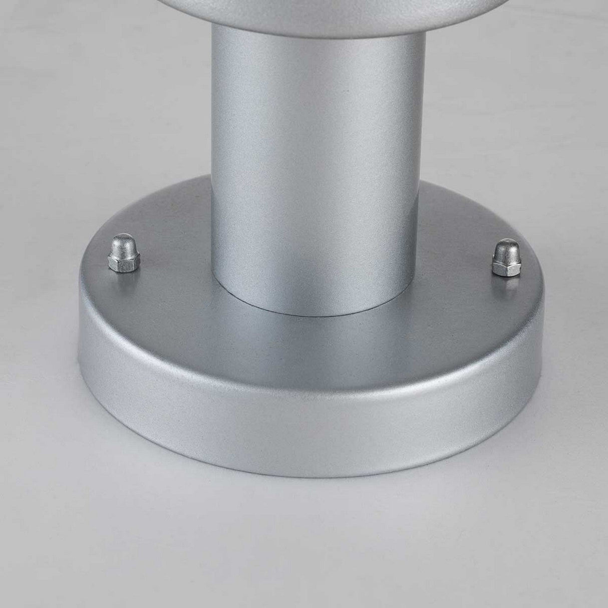 A close-up view of the Klampenborg 1 Light Pedestal in silver reveals its cylindrical, metallic base inspired by Scandinavian design, featuring two hexagonal bolts on opposite sides. It rests on a white surface with a smooth, matte finish that embodies the sleek style characteristic of the Klampenborg line.