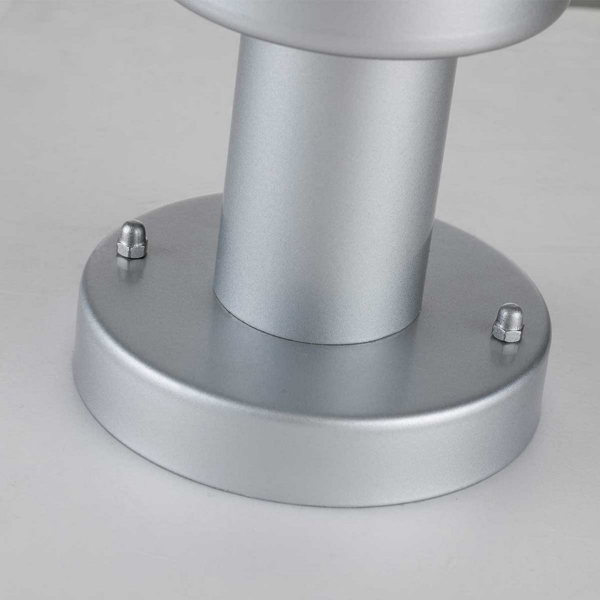 A close-up of the Klampenborg 1 Light Pedestal in silver, featuring a metallic cylindrical design reminiscent of sleek Scandinavian aesthetics, attached to a round base with two visible screws on a white surface.
