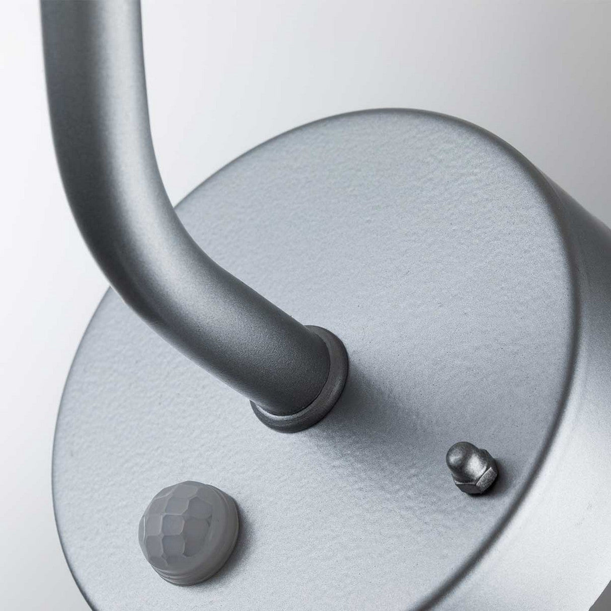 Detailed image of the Klampenborg 1 Light Outdoor Wall Light with PIR in Silver, showcasing its circular metallic design and curved handle at the top. Crafted from sleek stainless steel, it includes a small white PIR sensor and a metal protrusion on its surface. Set against a soft gray background, emphasizing its contemporary aesthetic.
