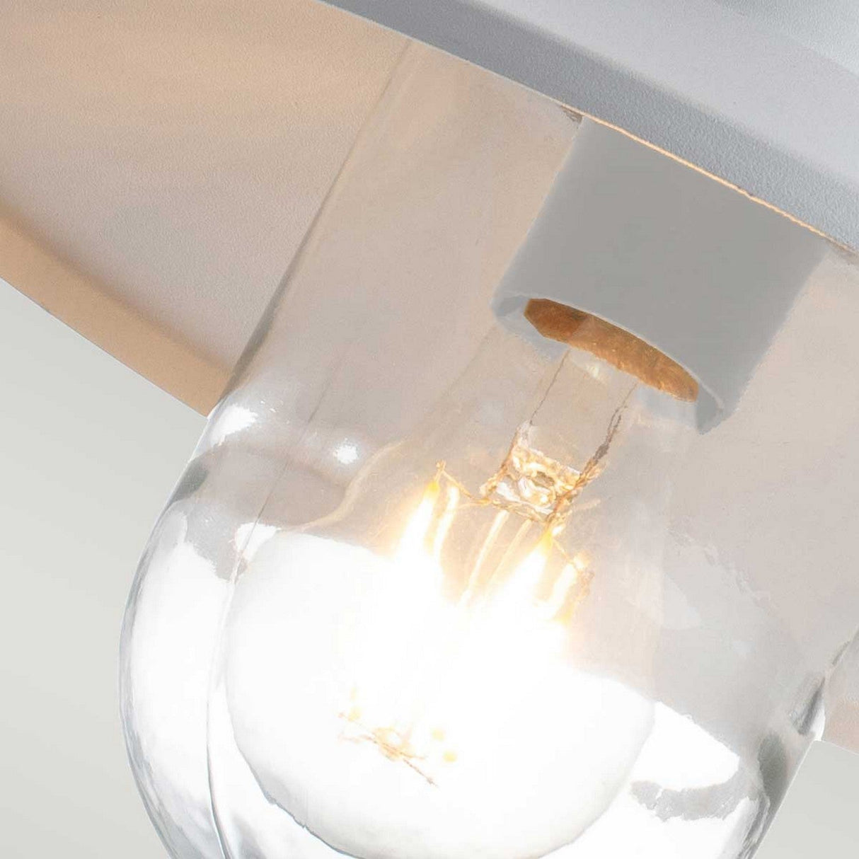 Close-up of the Klampenborg 1 Light Outdoor Wall Light with PIR in white, showcasing its incandescent bulb casting a warm glow reminiscent of Scandinavian design, gently illuminating the surrounding area from its ceiling-mounted position inside a clear glass fixture.
