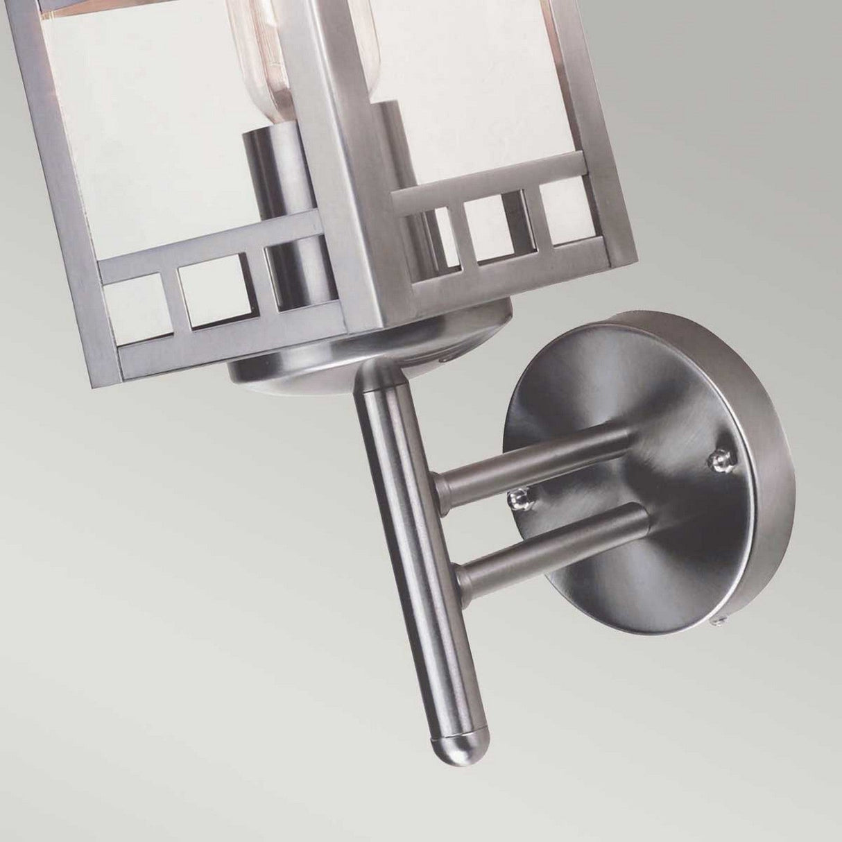 Featuring a close-up of the Kolne 1 Light Outdoor Wall Light in a Scandinavian design, this wall sconce exudes geometric flair. It showcases a rectangular frame and cylindrical socket set on a round base, all finished in brushed stainless steel. Positioned against a peaceful gray backdrop, it encapsulates sleek modern elegance.