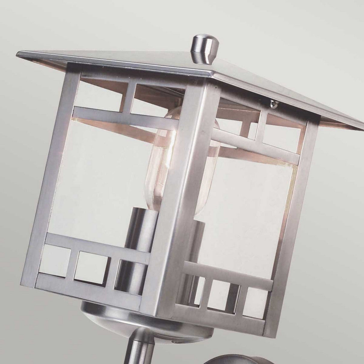 A detailed view of the Kolne 1 Light Outdoor Wall Light in stainless steel highlights its Scandinavian design, featuring a geometric pattern. The clear glass panels expose the bulb inside, and the square, peaked roof accompanied by a sleek finish enhances its stylish appearance.
