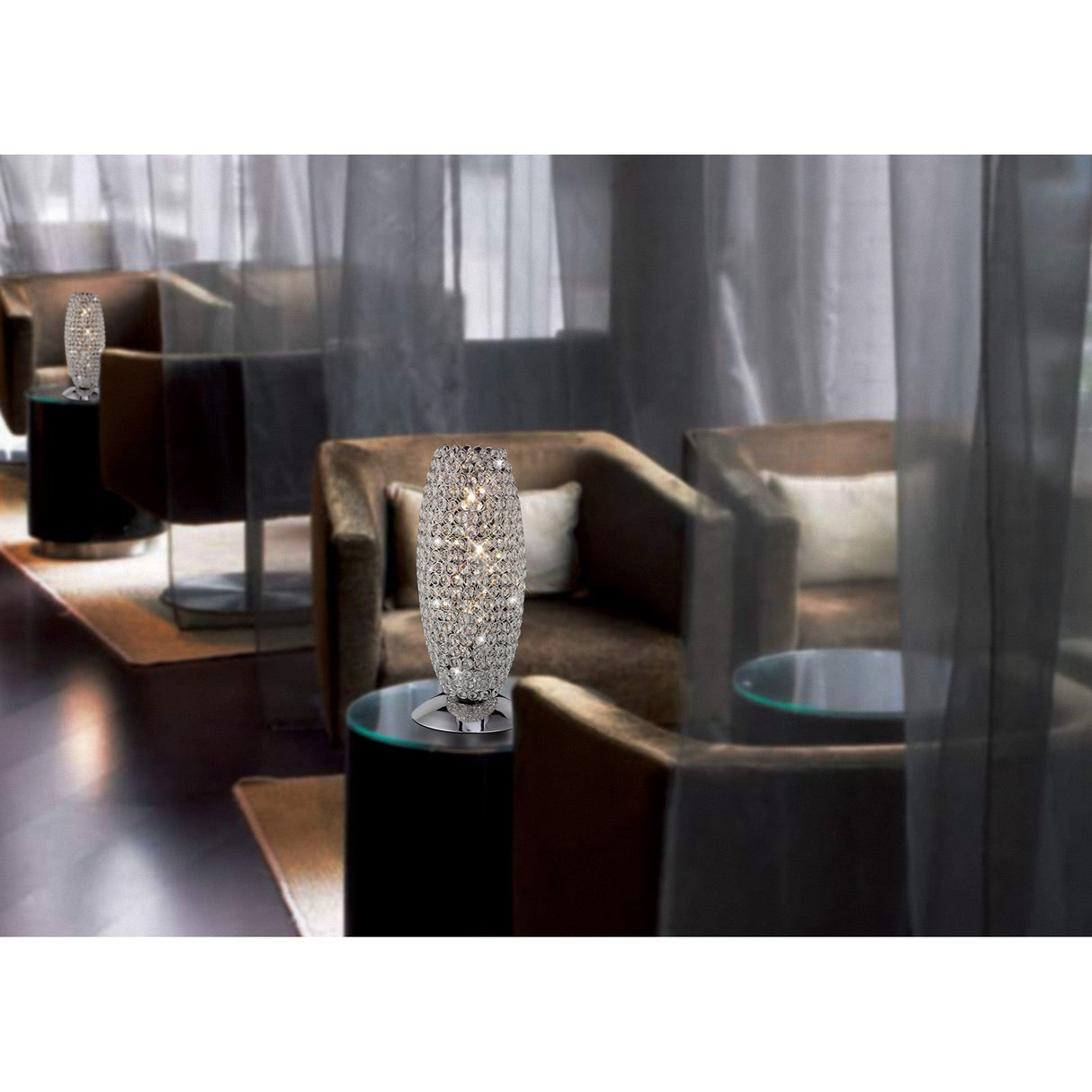 Elegant lounge area with brown armchairs, round glass-topped tables, and Kos 3 Light Crystal Table Lamps in polished chrome. Sheer gray curtains in the background enhance the modern, sophisticated vibe.