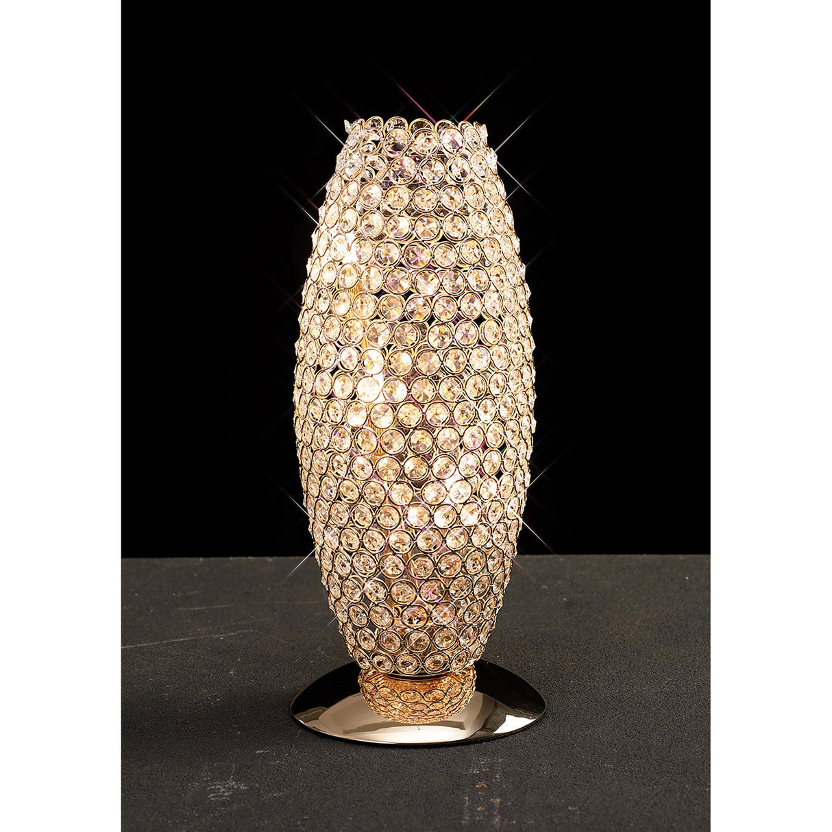 The Kos 3 Light Crystal Table Lamp in French Gold features a tall, cylindrical shape adorned with gold-colored crystals on a metallic base, adding luxurious decorative lighting and brilliance against a black background.