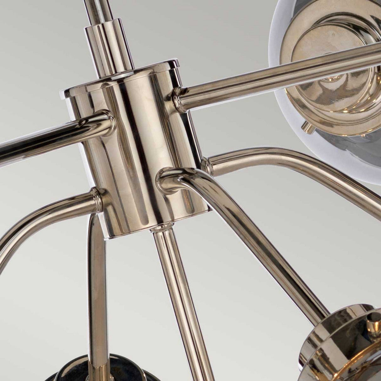 The Kula 9 Light Chandelier - Polished Nickel features a close-up view of its multiple arms extending outward. Its polished nickel finish elegantly reflects light, accentuating the intricate connection points, while its design brings a touch of mid-century modern style against the soft, neutral gray background.