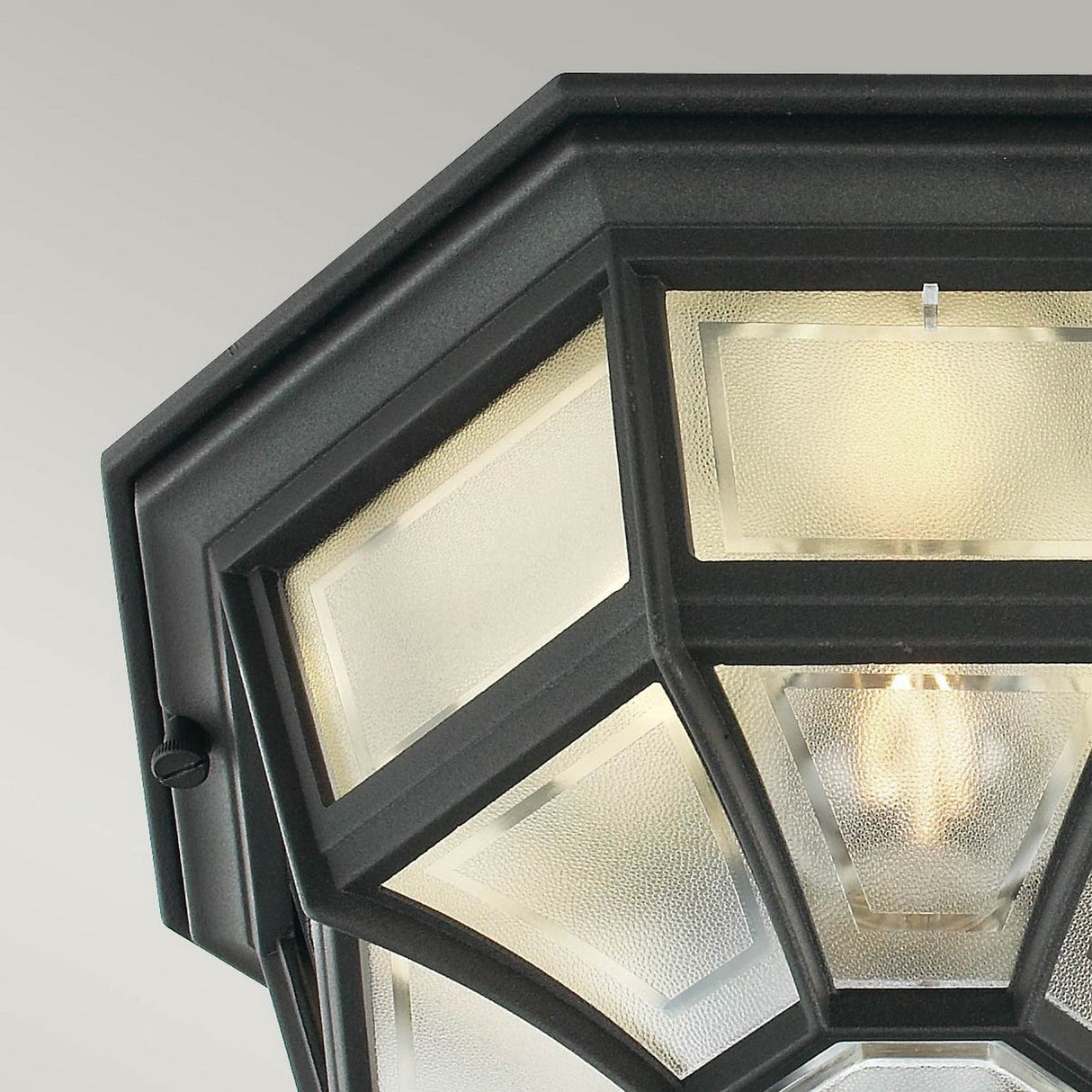 The Latina Outdoor 1 Light Ceiling Light in black is showcased in a close-up, highlighting its geometric design and frosted glass panels that exude elegance with a black powder coating. The illuminated bulb emits a warm glow, beautifully contrasting against the light gray background, making it an ideal choice for any space requiring an IP44-rated fixture.