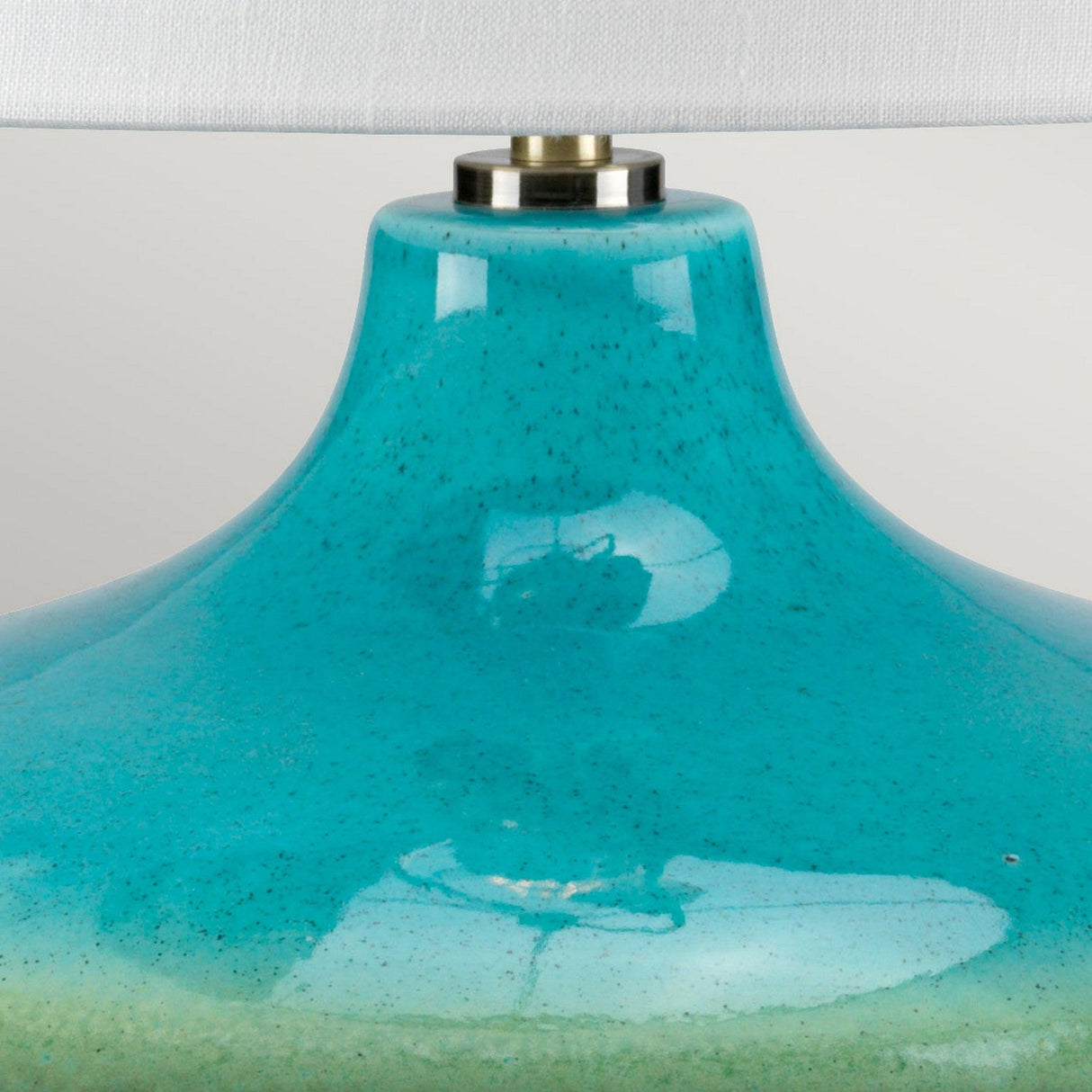A close-up of the Laguna 1 Light Small Table Lamp reveals its turquoise and graphite glaze ceramic base, featuring a slightly speckled, reactive surface with a smooth, glossy finish. A small metallic detail is present at the top, while part of the white lampshade can be seen above.