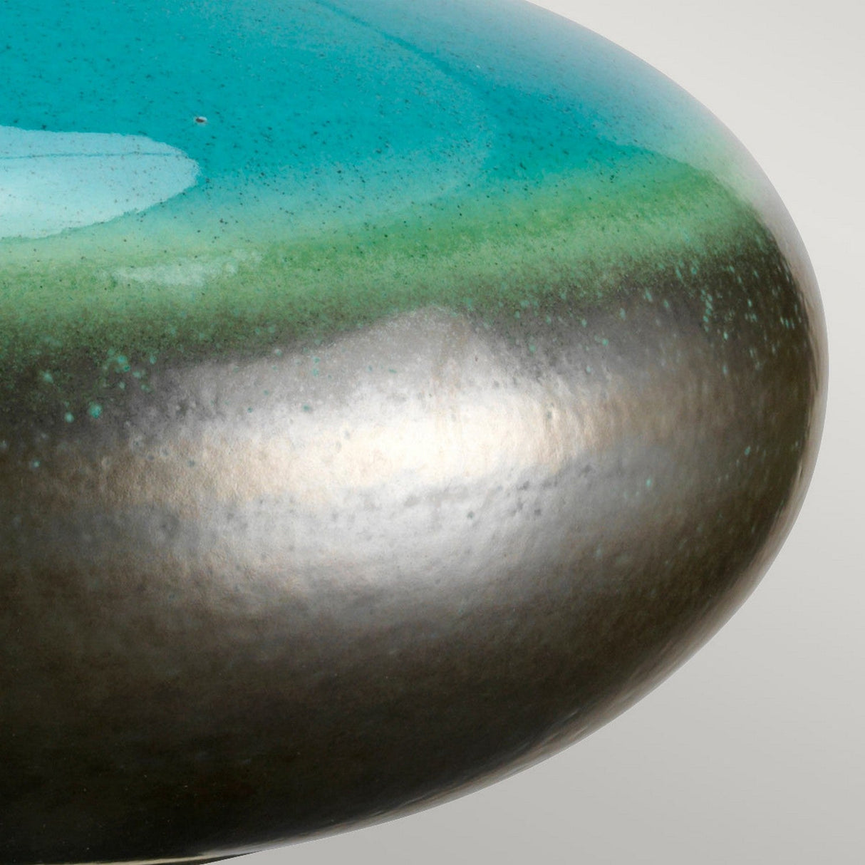 Close-up of a Laguna 1 Light Small Table Lamp - Turquoise & Graphite Glaze, showcasing its smooth, rounded surface. It displays a gradient of colors from turquoise to graphite to dark metallic, accentuated by a reactive glaze that provides a glossy, reflective finish. The background is softly set in neutral gray.