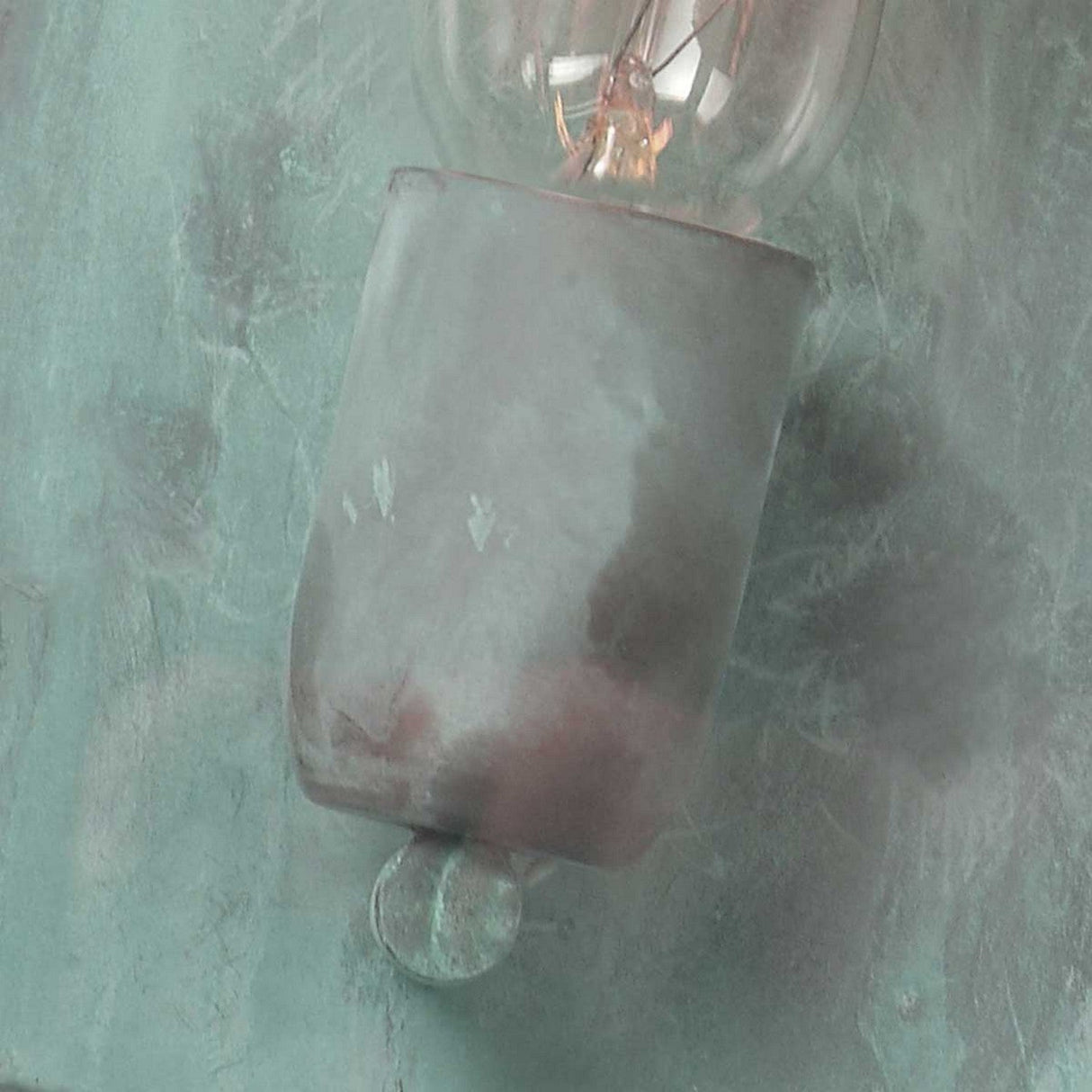 Against a textured, muted green background with subtle brush strokes, a frosted glass light bulb captures the timeless allure of the Lambeth Palace 1 Light Outdoor Wall Light in Verdigris. The bulb’s slightly worn appearance adds to its rustic charm, reminiscent of this classic outdoor lighting piece.