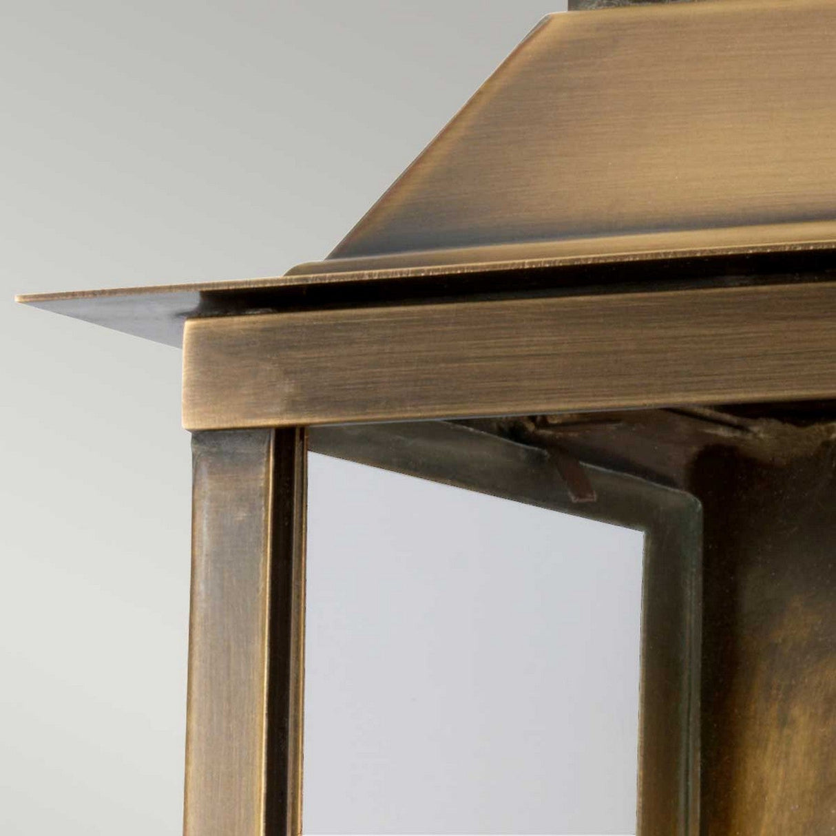 A close-up of the Lambeth Palace 1 Light Outdoor Wall Light reveals its vintage-style design with clear glass panels. The aged brass finish offers a slightly weathered look that adds rustic charm to the piece. Set against a plain, light gray backdrop, it radiates timeless elegance.