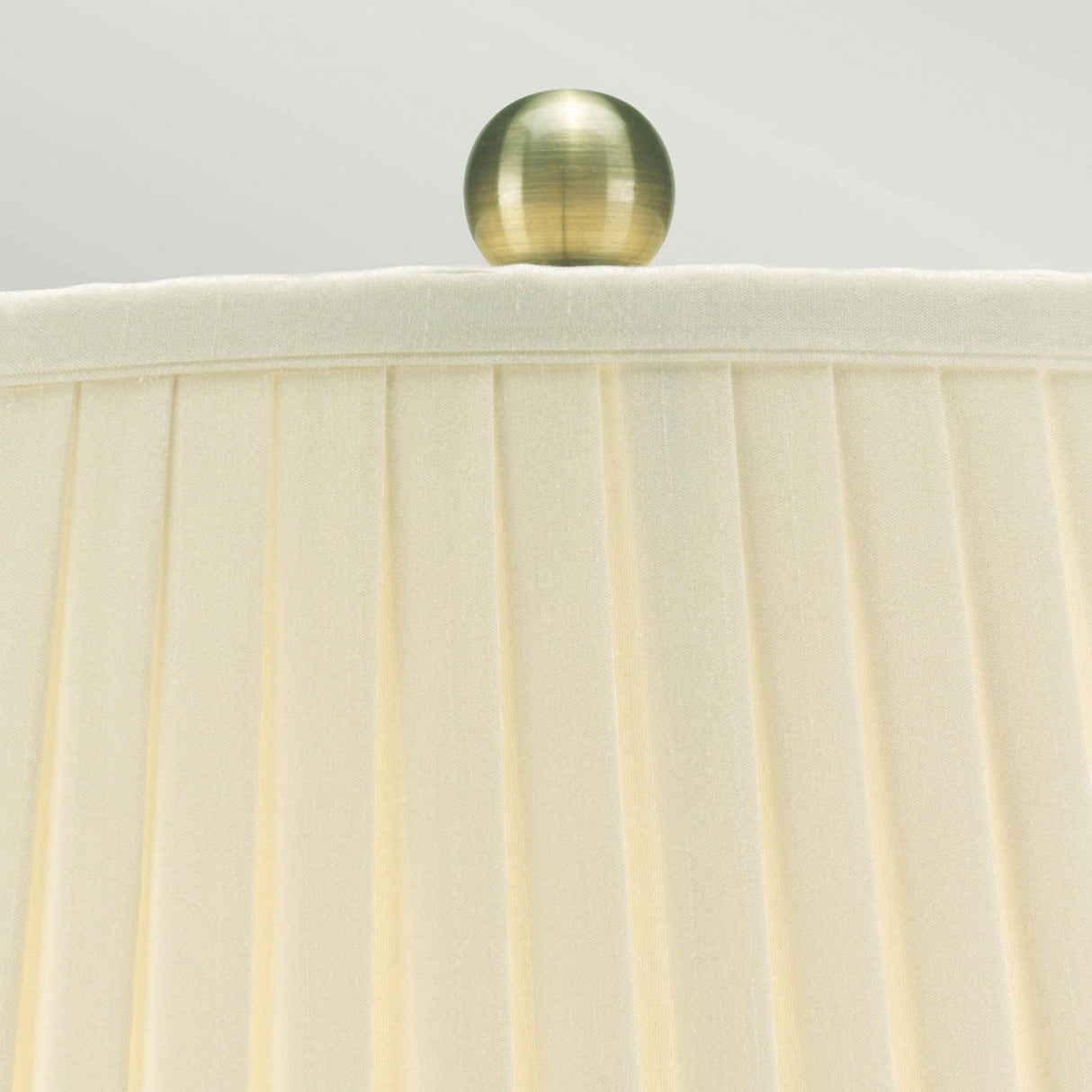 A close-up view of the Leaves Brown Gold 1 Light Table Lamp, featuring an ivory pleated fabric lampshade accented with a brass-colored spherical finial. The elegant design is set against a soft gradient background of off-white and light gray, evoking the delicate richness of fine Chinese porcelain.
