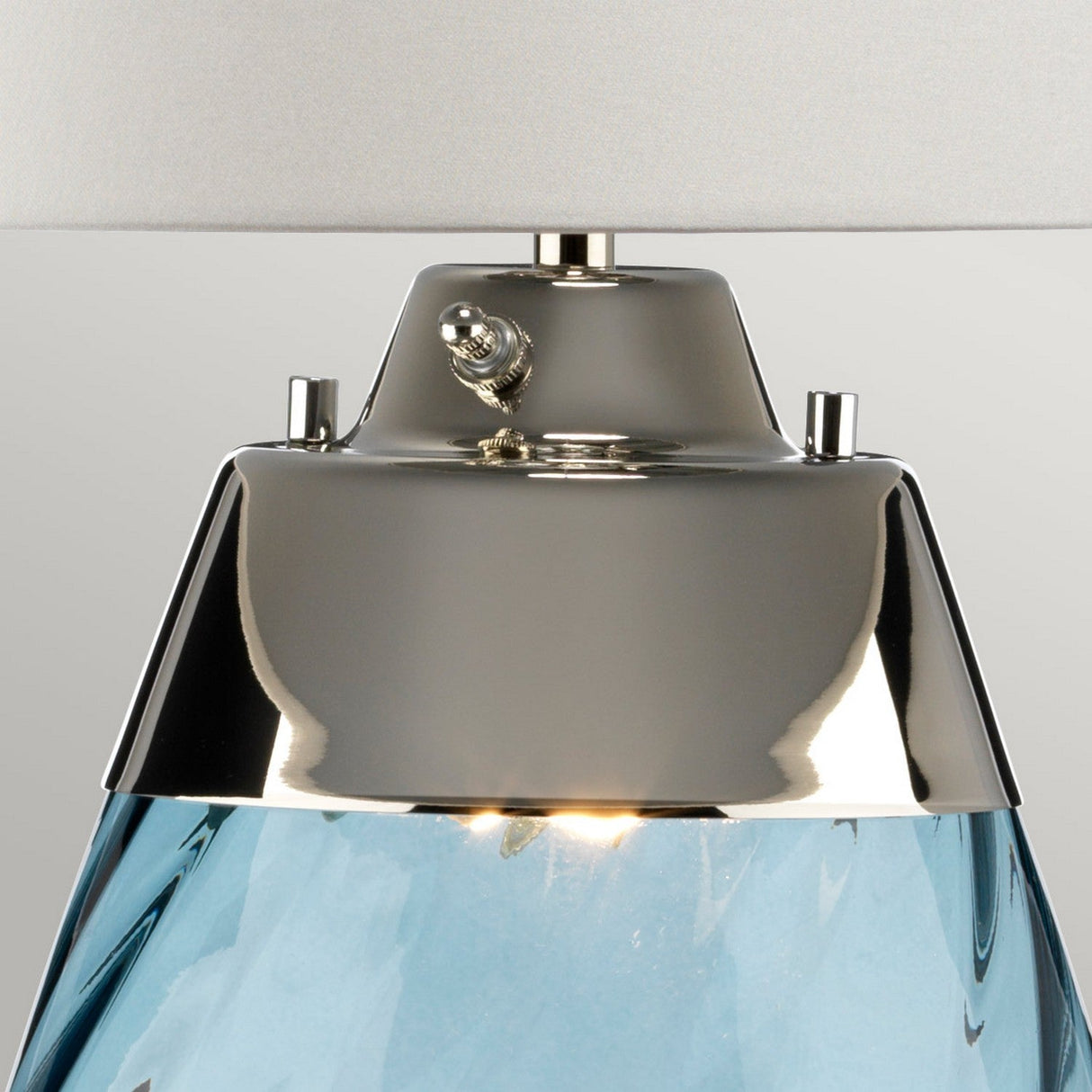 Close-up of the Lena 2 Light Large Blue Table Lamp, featuring a metallic top with a small toggle switch. This lamp showcases a mouth-blown blue glass base that elegantly reflects light. Its off-white shade complements the design, while the dual-light feature adds versatility to its modern charm.