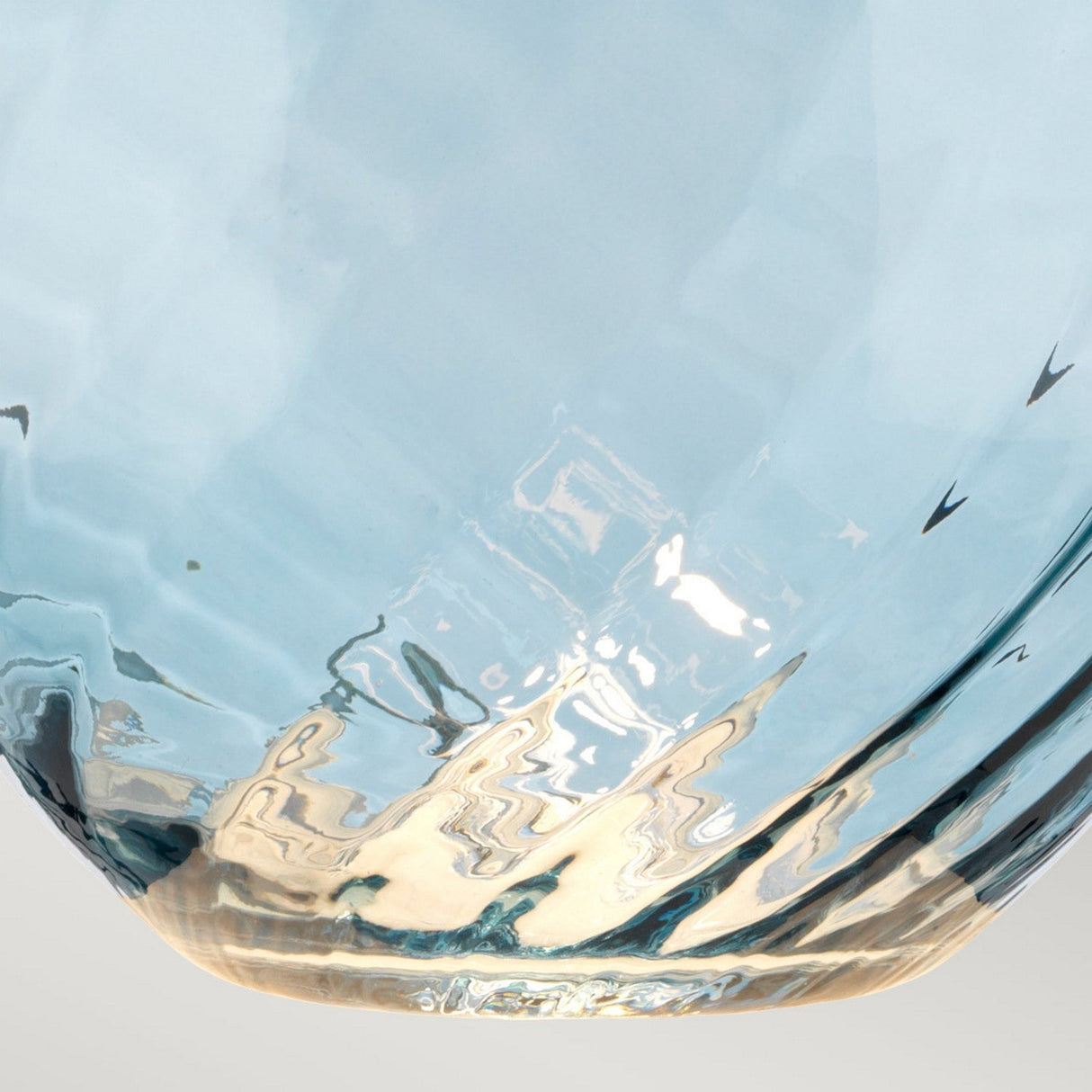 Close-up of the mouth-blown blue glass base on the Lena 2 Light Large Blue Table Lamp - Off-White Shade, showcasing its dual-light feature that casts a gentle glow and creates abstract patterns across its transparent surface. Reflective highlights dance on its curved shape against a neutral background, echoing the elegance of the lamp design.