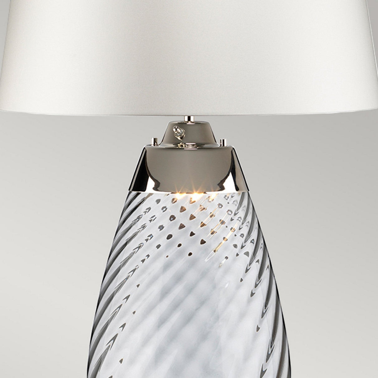 The Lena 2 Light Small Blue Table Lamp features a twisted, mouth-blown glass base with an elegant metal accent, all complemented by an off-white shade. This stylish table lamp offers a modern look while softly illuminating the intricate design of the glass.