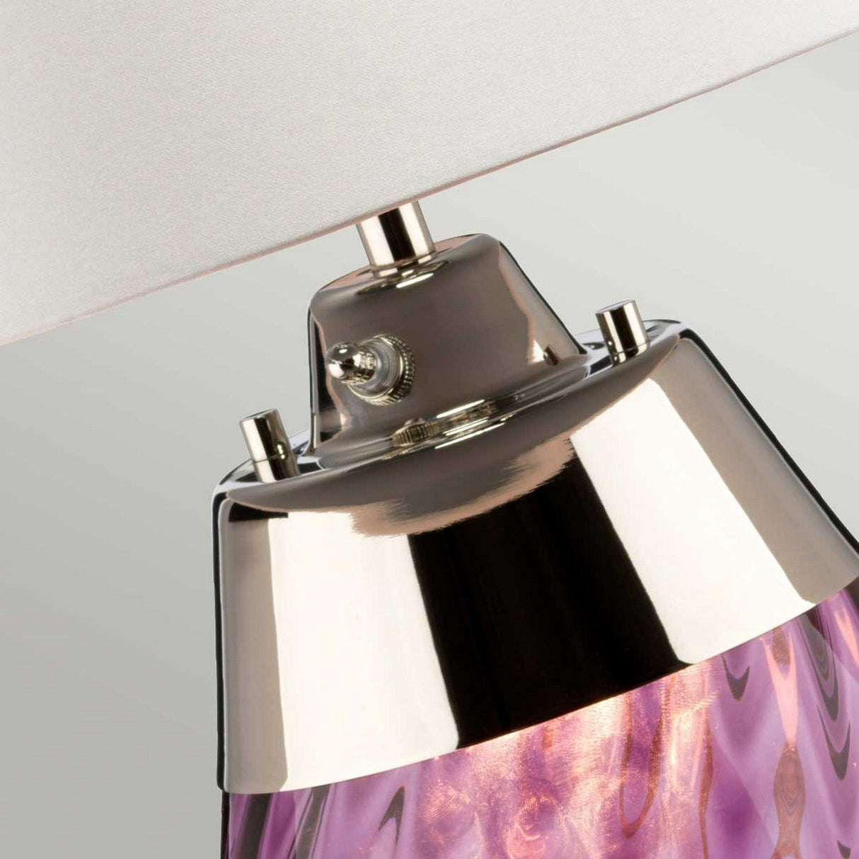 Close-up of the Lena 2 Light Small Plum Table Lamp featuring a glossy silver base and a richly textured purple body. The switch is located on the silver part beneath the off-white shade. This lamp boasts a dual-lit system, emanating a warm glow through its beautiful purple glass.
