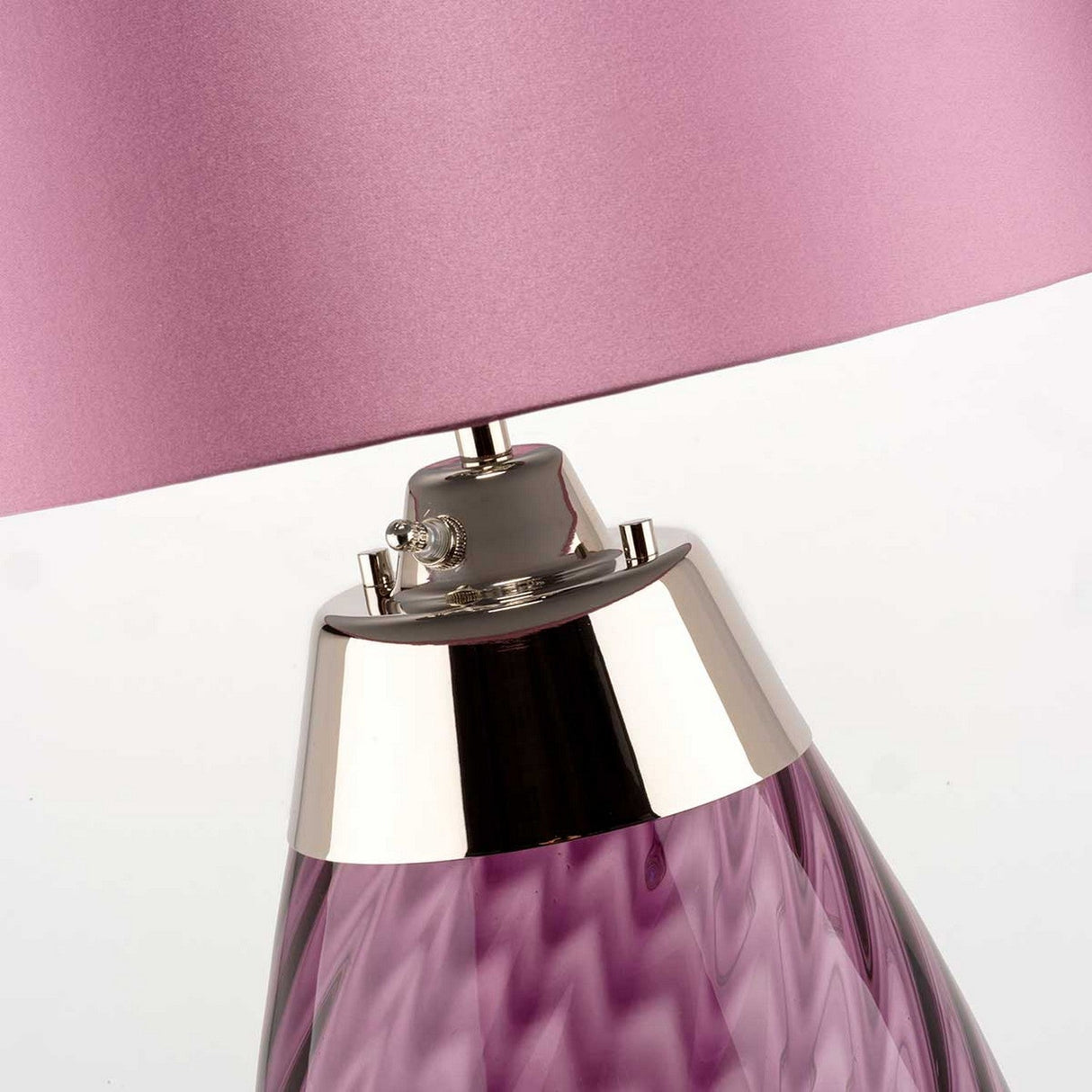 A close-up of the Lena 2 Light Small Plum Table Lamp highlights its textured base and metallic stand. The lampshade, crafted from heather satin, matches in a soft pinkish-purple hue and appears slightly tilted. The switch for the Dual-Lit system is visible on the metallic section.