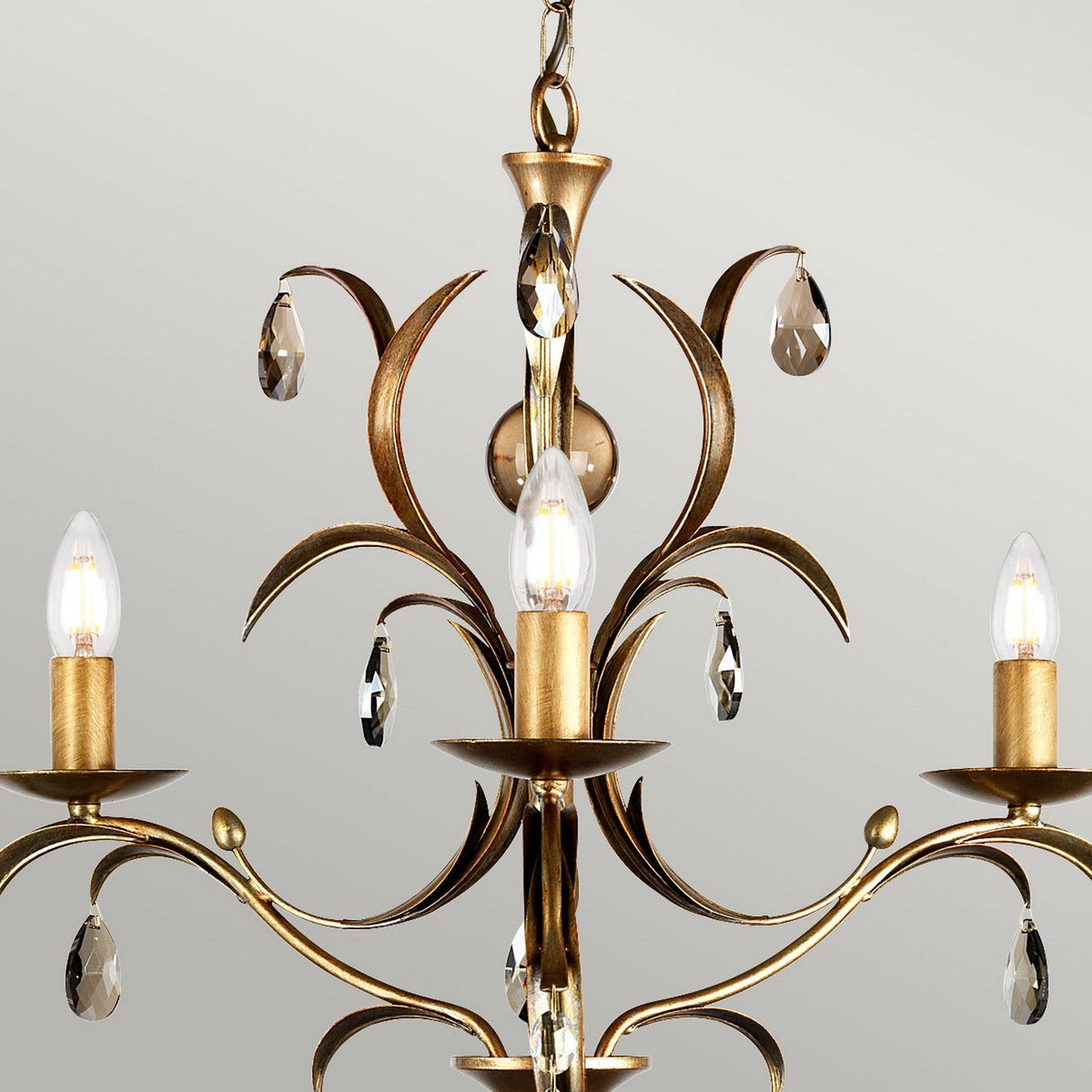 The Lily 3 Light Chandelier - Bronze Patina features an elegant floral design with a hand-patinated bronze finish. It includes three candle-shaped light bulbs and smoked cut glass droplets, all beautifully contrasted against a light gray background for a sophisticated look.