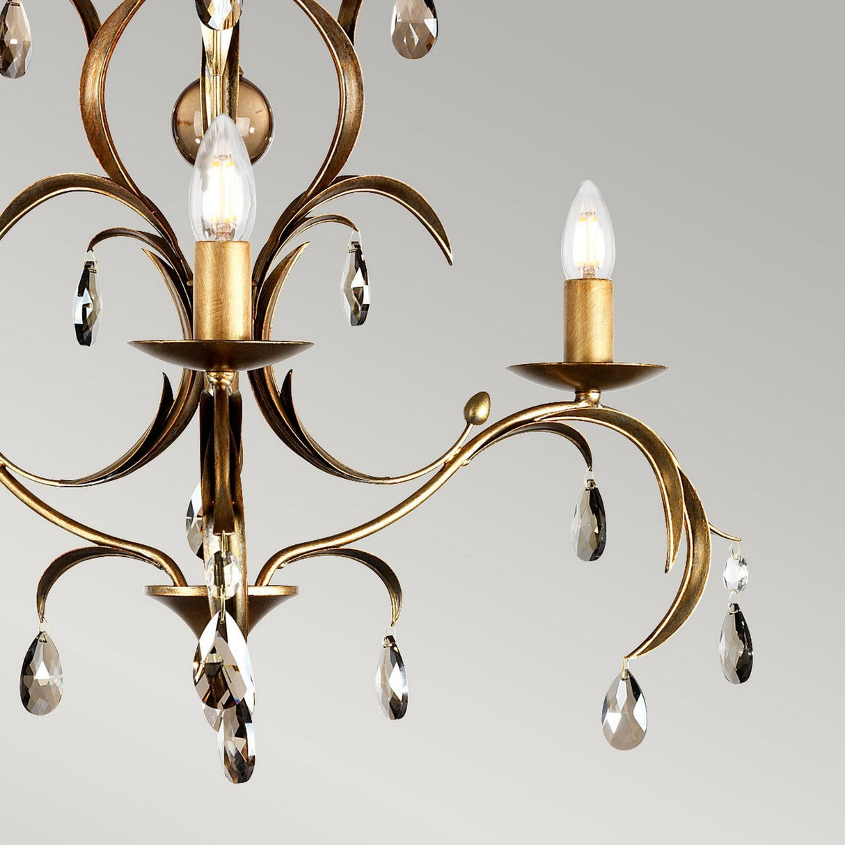 Close-up of the Lily 3 Light Chandelier - Bronze Patina, showcasing two glowing candle-like bulbs. The intricate design features gracefully curved metal arms adorned with smoked cut glass droplets and dangling crystal pendants, all set in a hand-patinated bronze finish against a light gray backdrop.