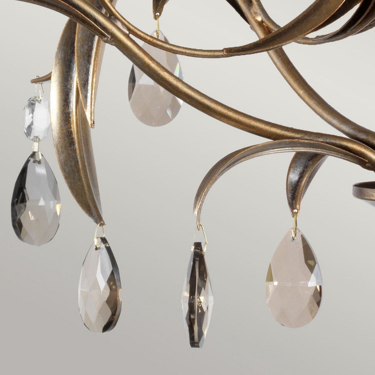 The Lily 5 Light Chandelier - Bronze Patina features a close-up view of teardrop-shaped, smoked cut glass droplets elegantly suspended from gold, leaf-like metal branches. With its bronze finish set against a neutral background, the crystals beautifully reflect light to create an elegant and luxurious appearance.