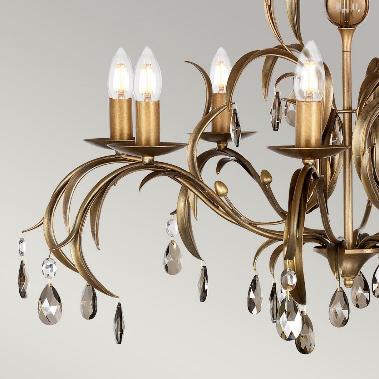 A close-up of the Lily 8 Light Chandelier - Bronze Patina highlights its gold and bronze finish with candle-like bulbs and intricately curved branches, while smoked cut glass pendants provide an elegant touch against a neutral background.